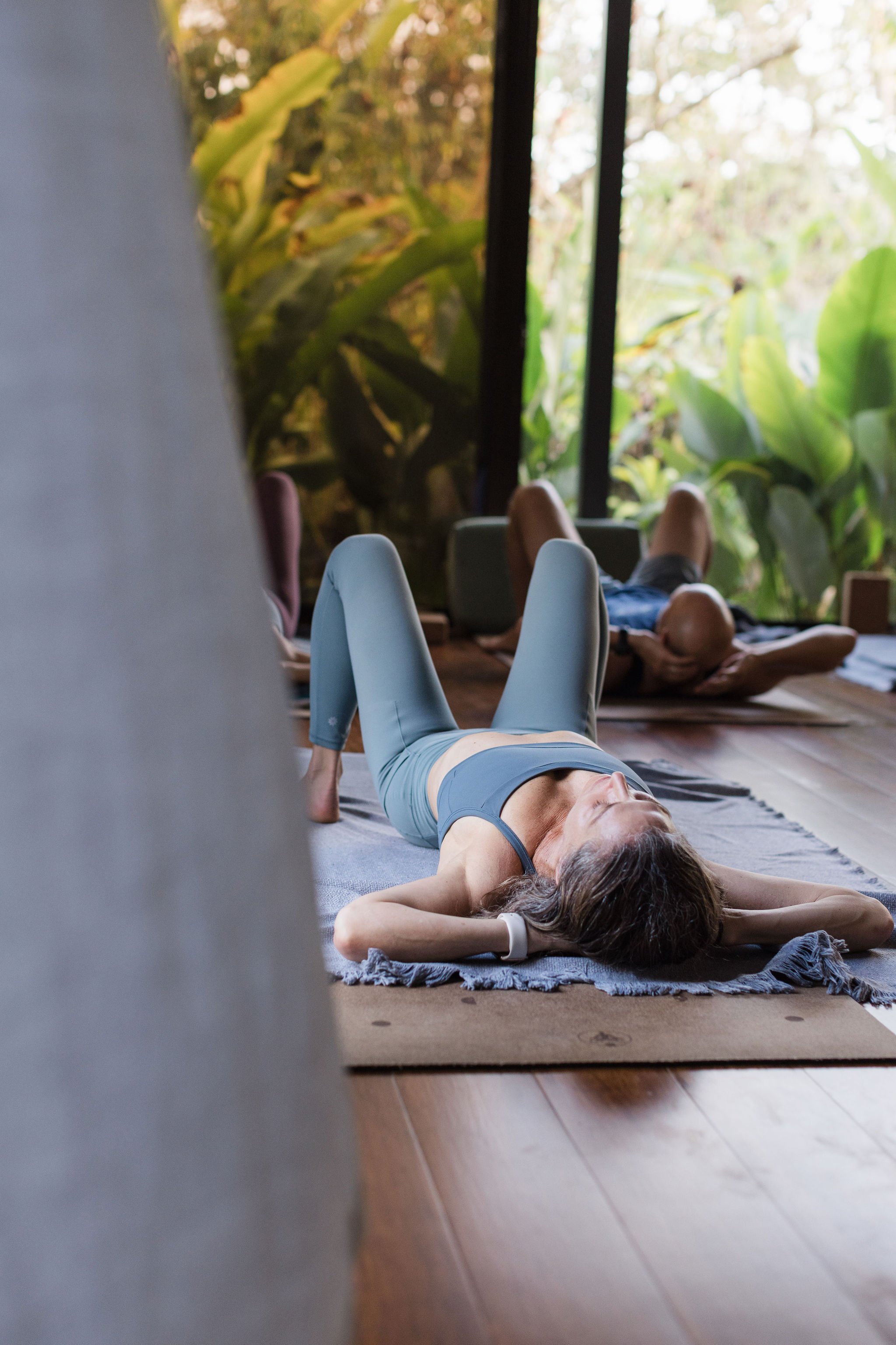 A woman practicing yoga in a peaceful room with large windows and lush greenery outside.