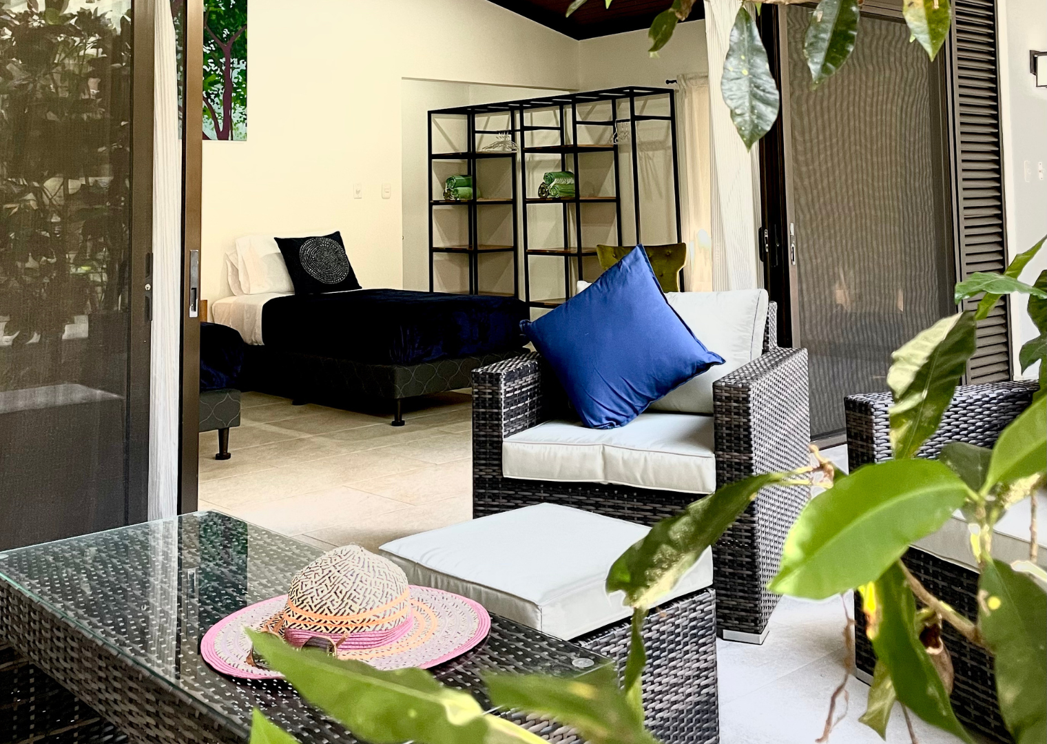 Interior of a living space with a bed, wicker armchairs with pillows, a glass table with a colorful hat, a shelving unit, and sliding glass doors with curtains, surrounded by green plants.