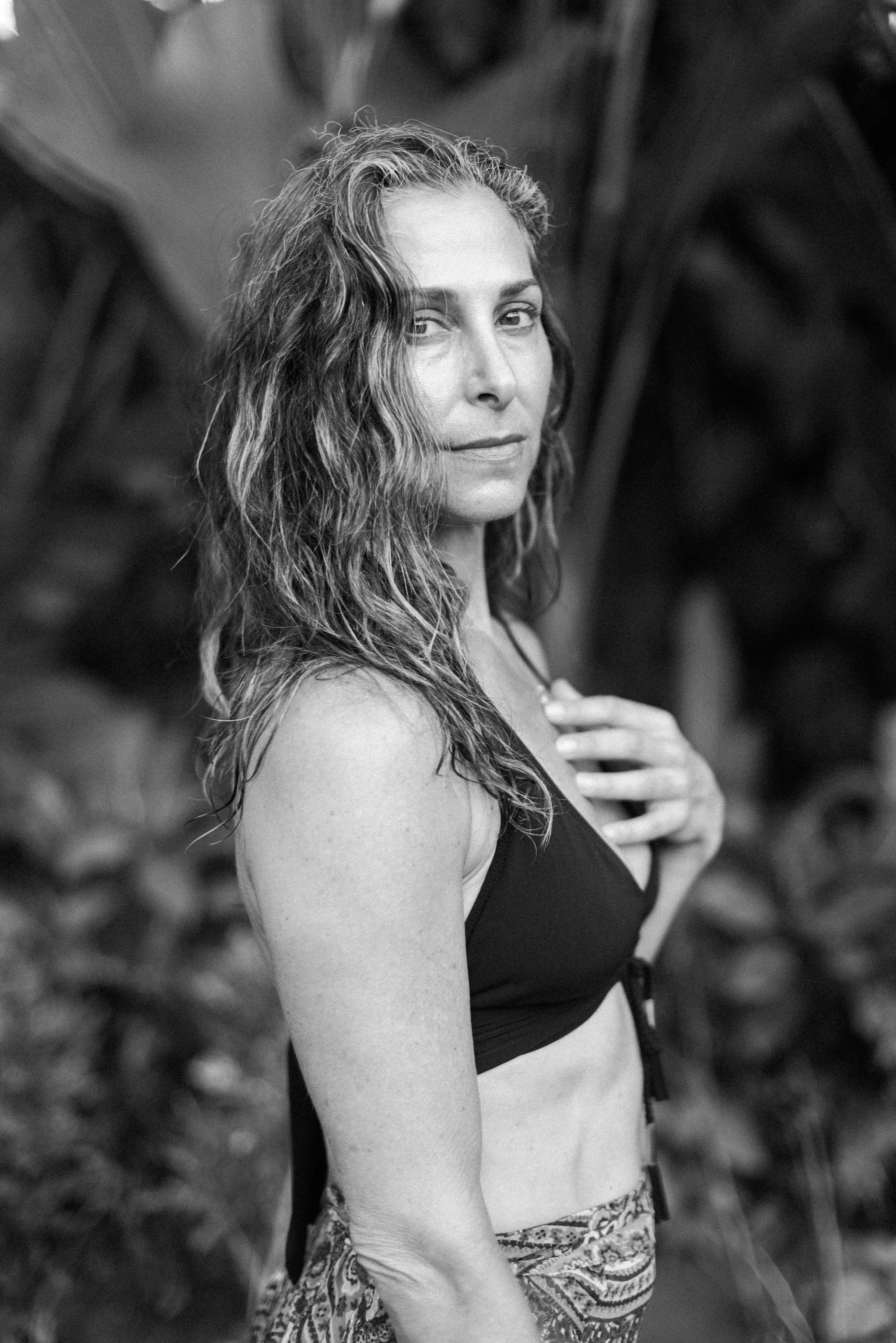A woman with wet, curly hair wearing a black bikini top and patterned shorts, standing outdoors with large leaves in the background.