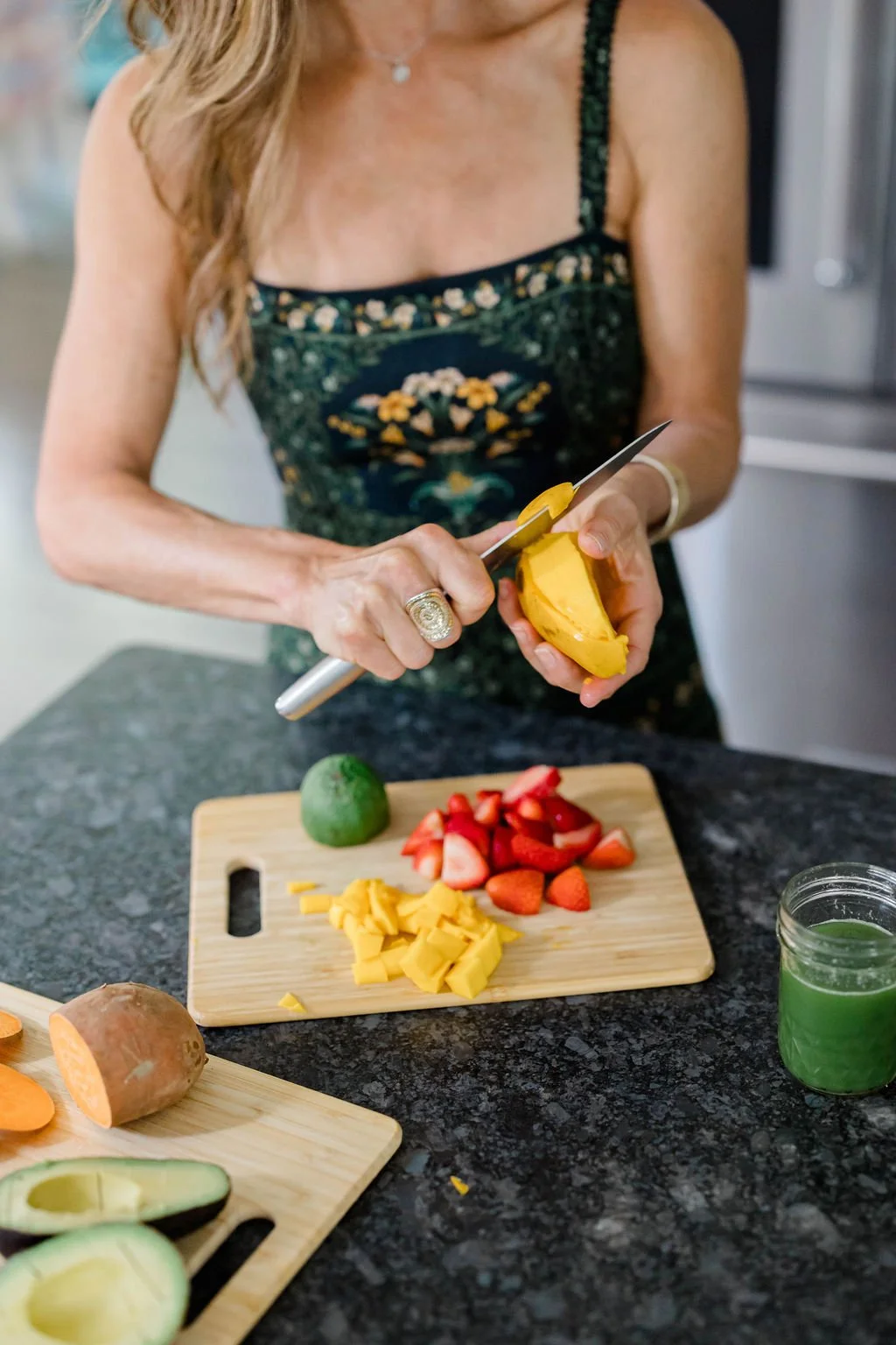 A woman in a floral dress is chopping a yellow mango on a kitchen counter. On the counter are cut strawberries, chopped yellow cheese, a whole mango, a sweet potato, and sliced avocados. There is also a small jar of green smoothie.