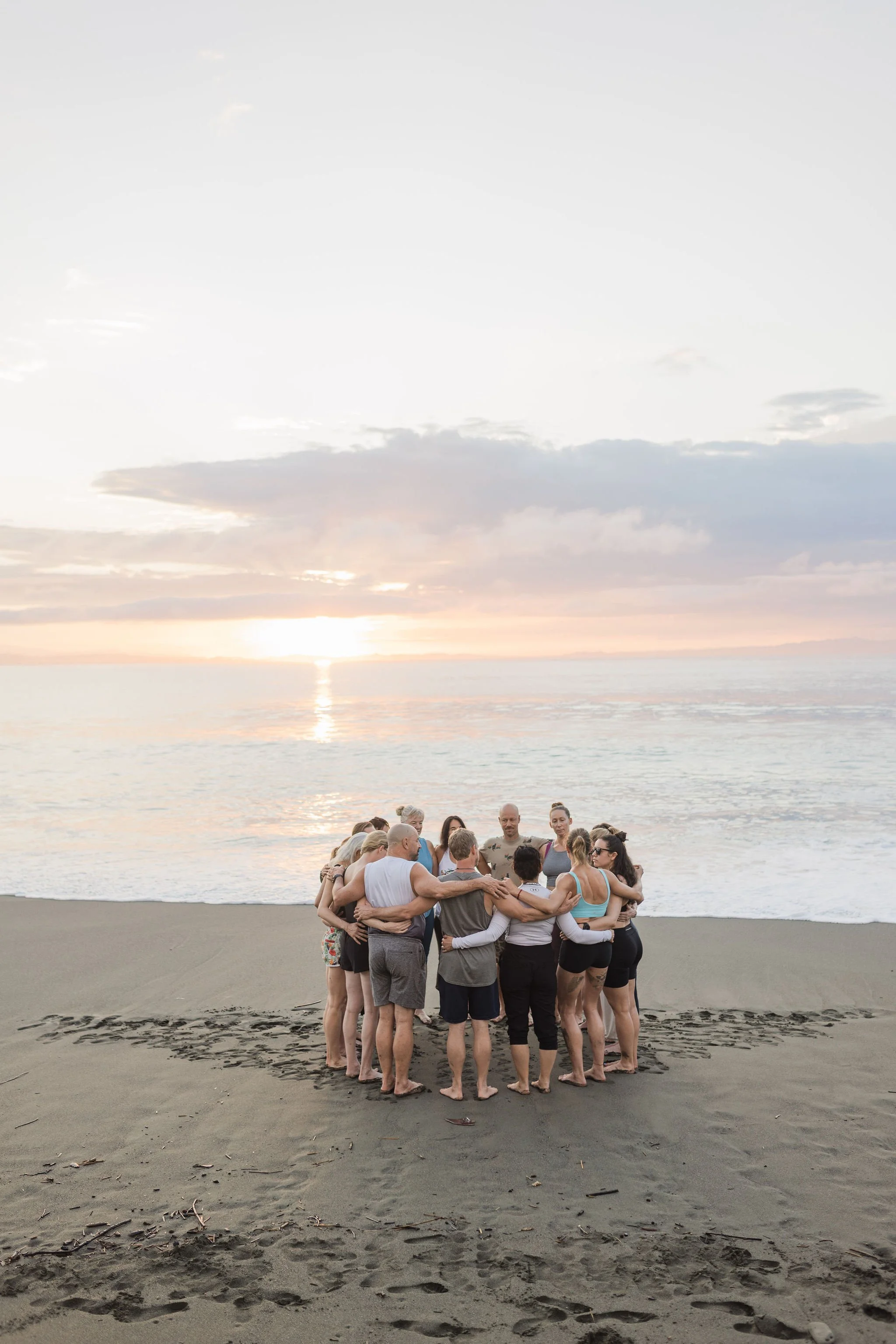 A group of people engaged in a circle huddle on a sandy beach with a calm ocean and a sunset sky in the background.