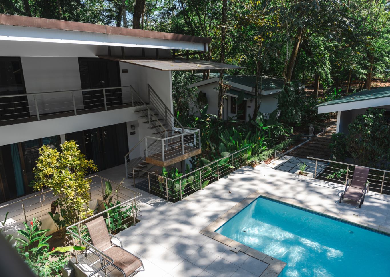 A modern house with a pool and a lush backyard, featuring white exterior walls, metal staircases, and surrounded by green trees and plants.