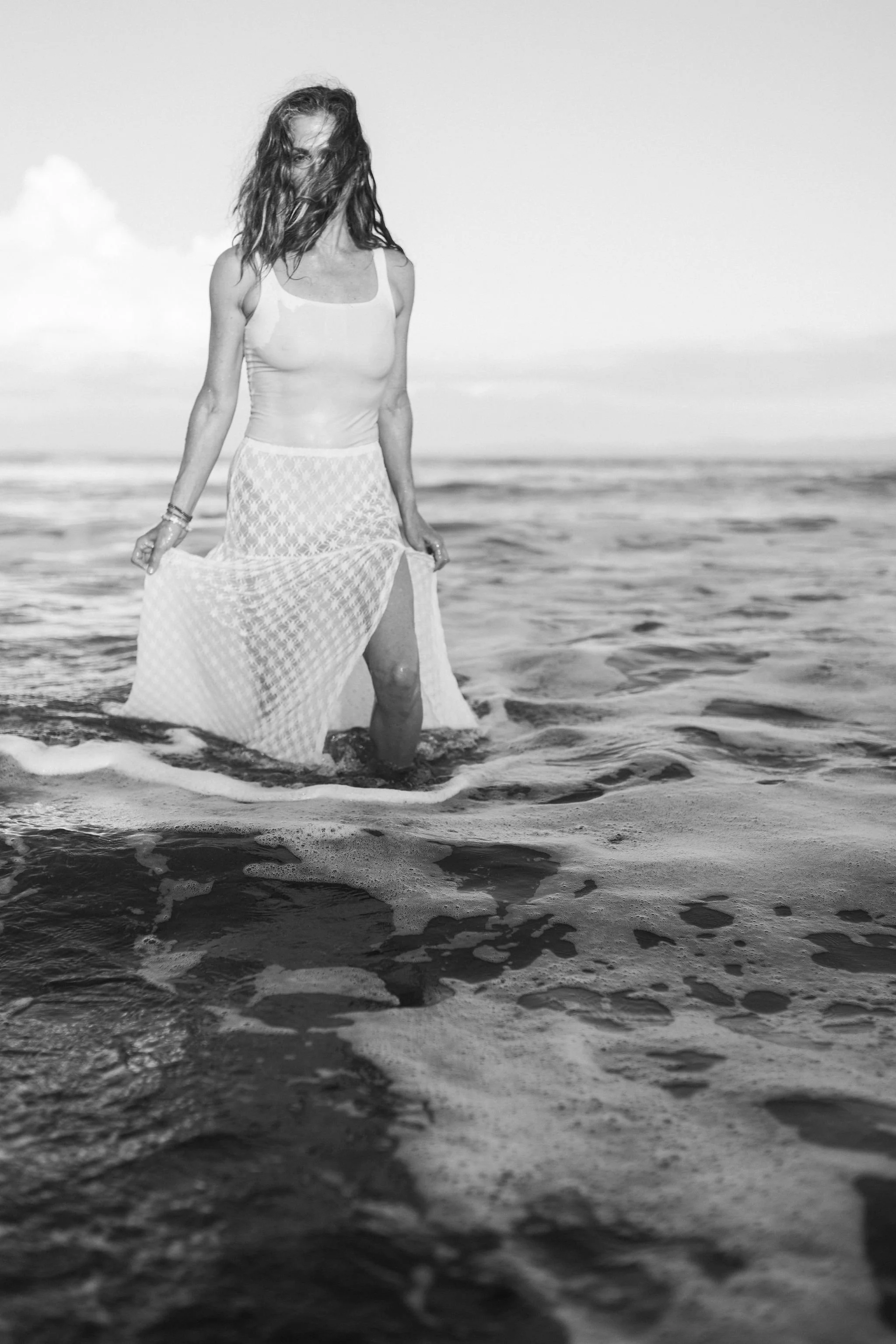 A woman standing in the ocean with her bare legs in the water, wearing a light-colored sleeveless top and a flowing, patterned skirt, with her hair blowing in the wind on a cloudy day.