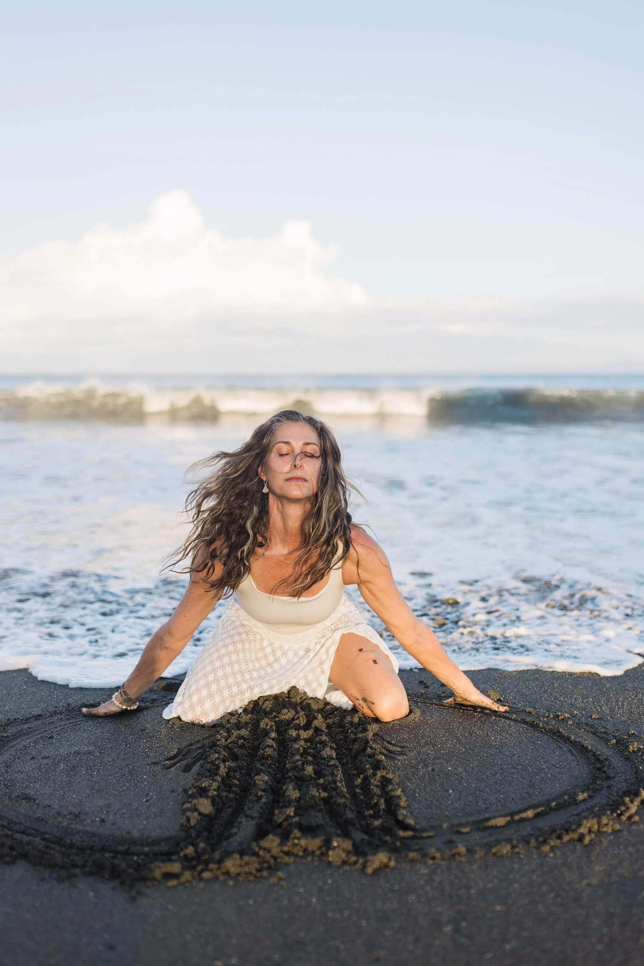 Woman with long hair sitting on a black sand beach, surrounded by a trail of sand, with ocean and cloudy sky in the background.