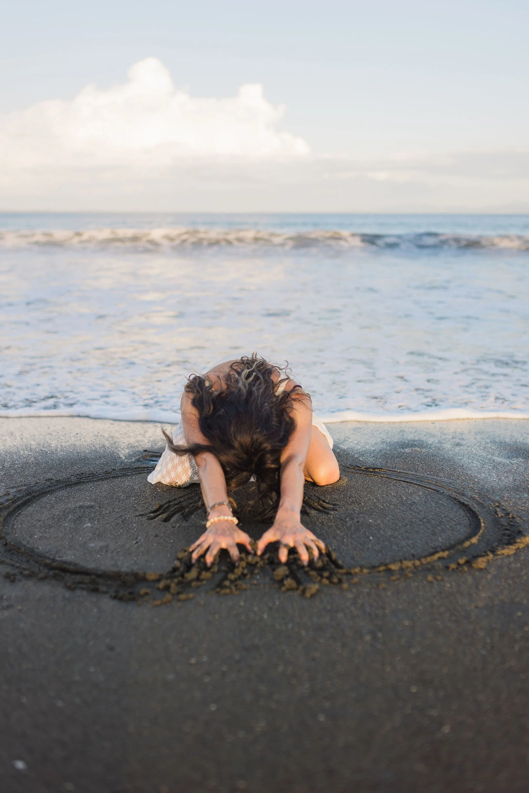 A person doing a yoga pose with arms extended forward on a sandy beach, with ocean waves and cloudy sky in the background.