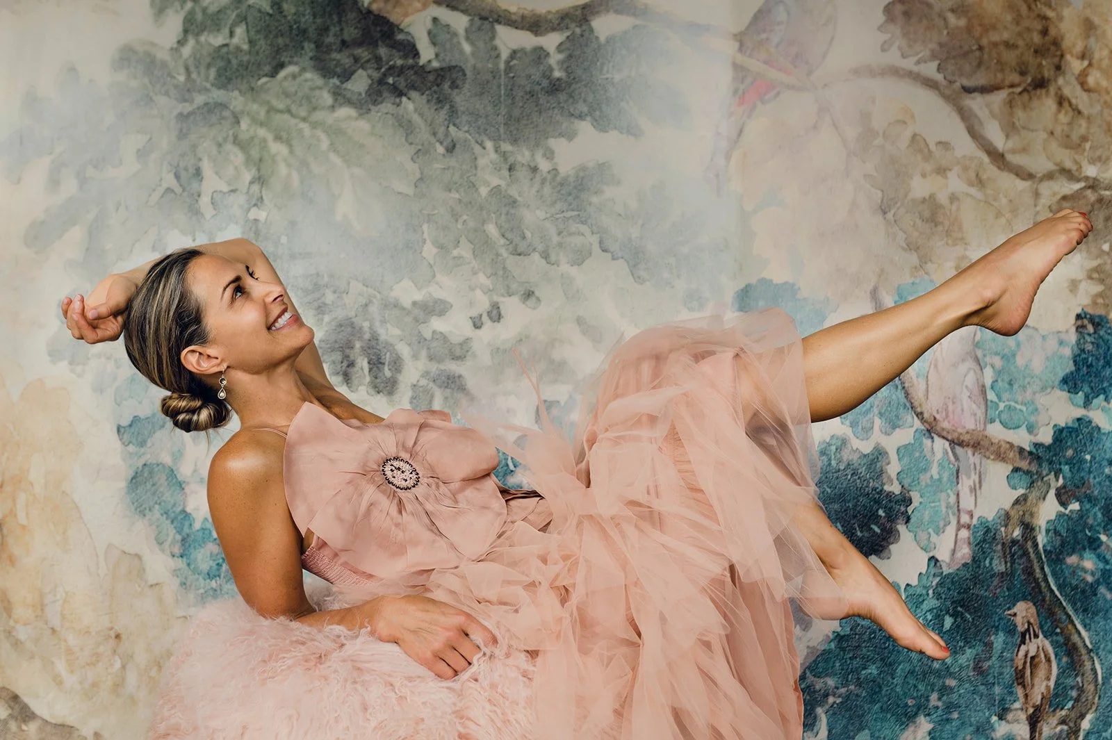 A woman in a peach-colored tulle dress is lying on a fluffy surface in front of a painted mural background, smiling and looking upwards with her left arm raised behind her head and her right arm resting on her stomach.