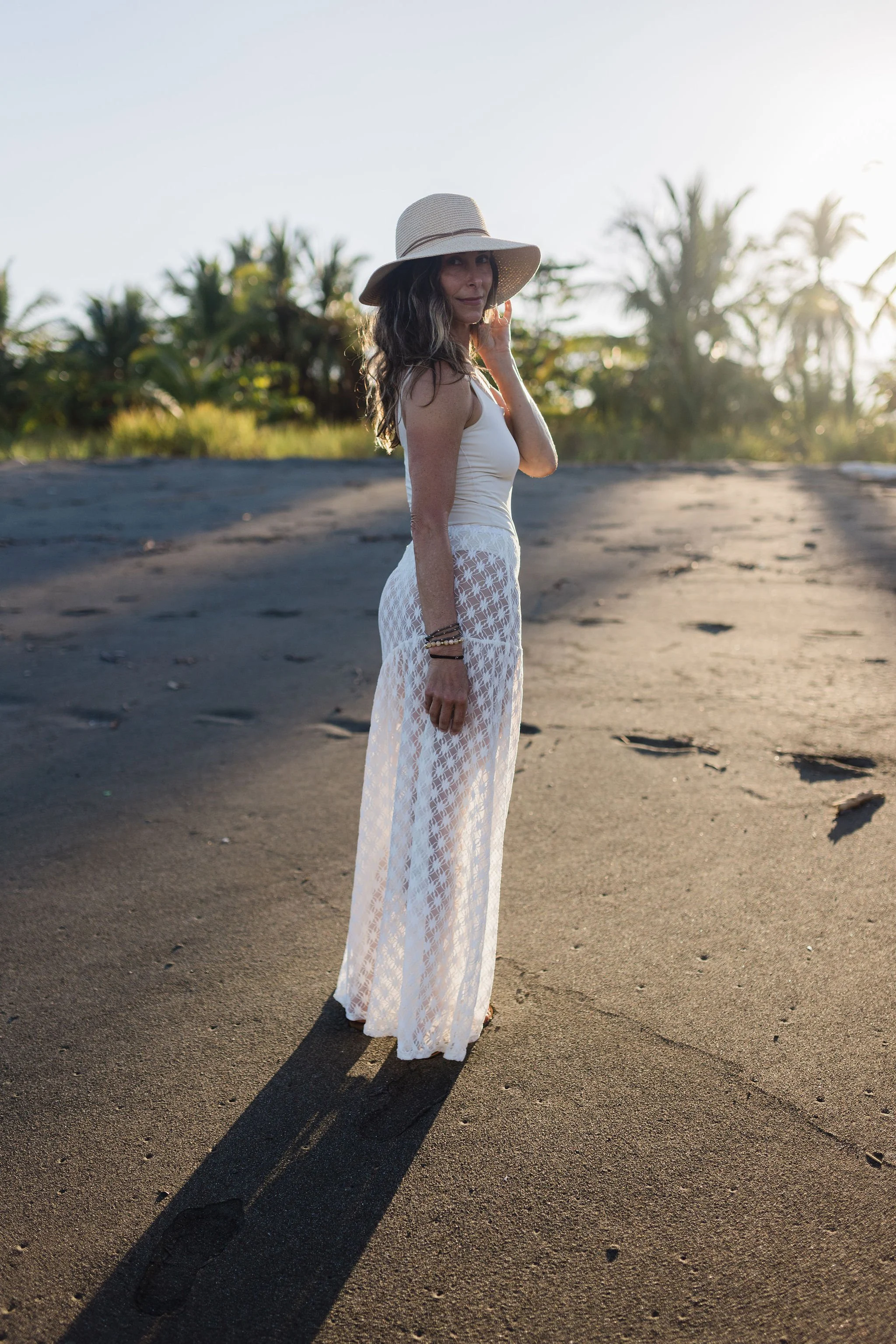 Lisa Dubaj wearing a wide-brimmed hat and white lace dress standing on sandy beach at a Costa Rica Resort enjoying the sunset.