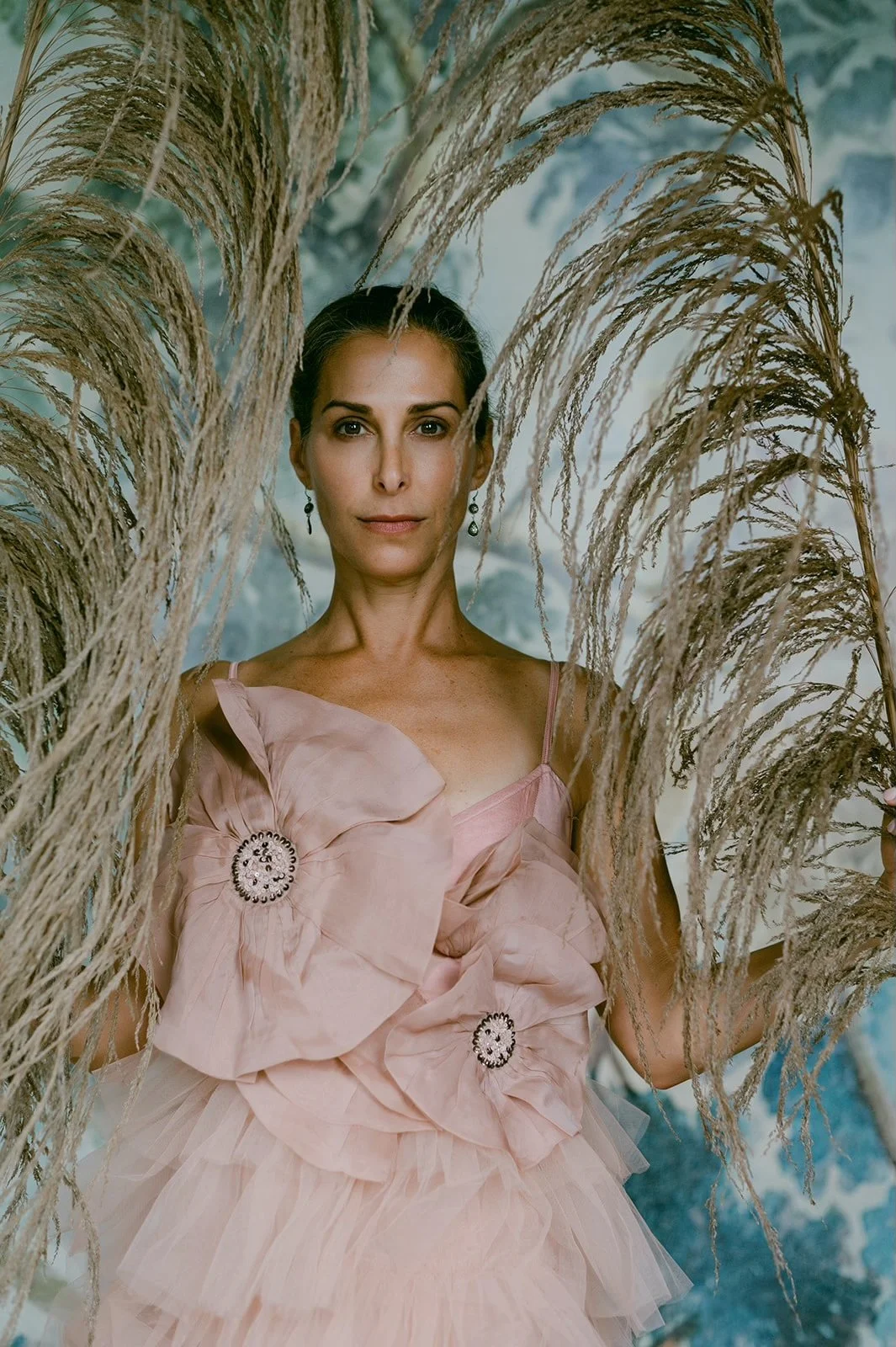 A woman wearing a pink dress with floral embellishments stands among tall, dried pampas grass.