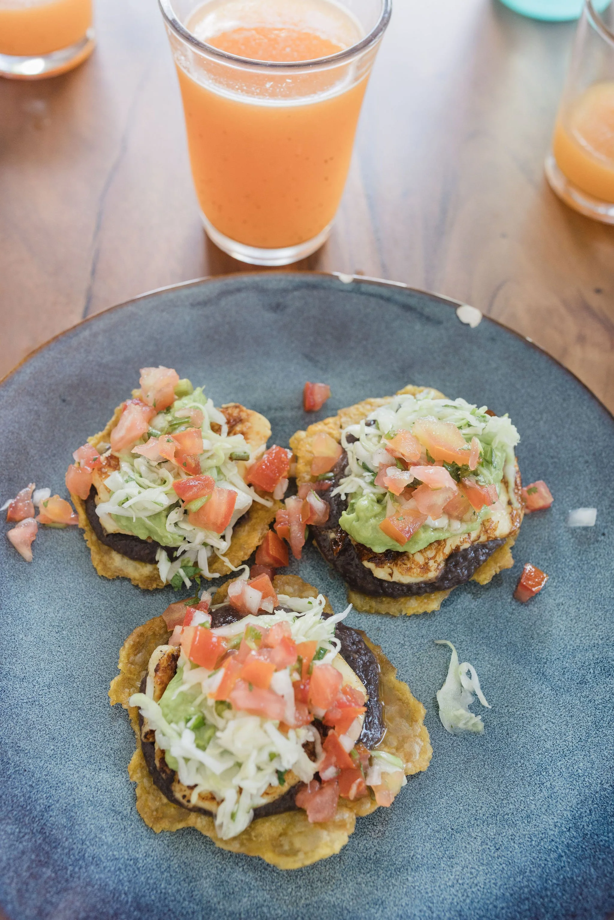 Close-up of three fried tacos topped with chopped tomatoes, lettuce, and shredded cheese on a gray plate. In the background, there is a glass of orange juice and additional glasses of orange beverage on a wooden table.