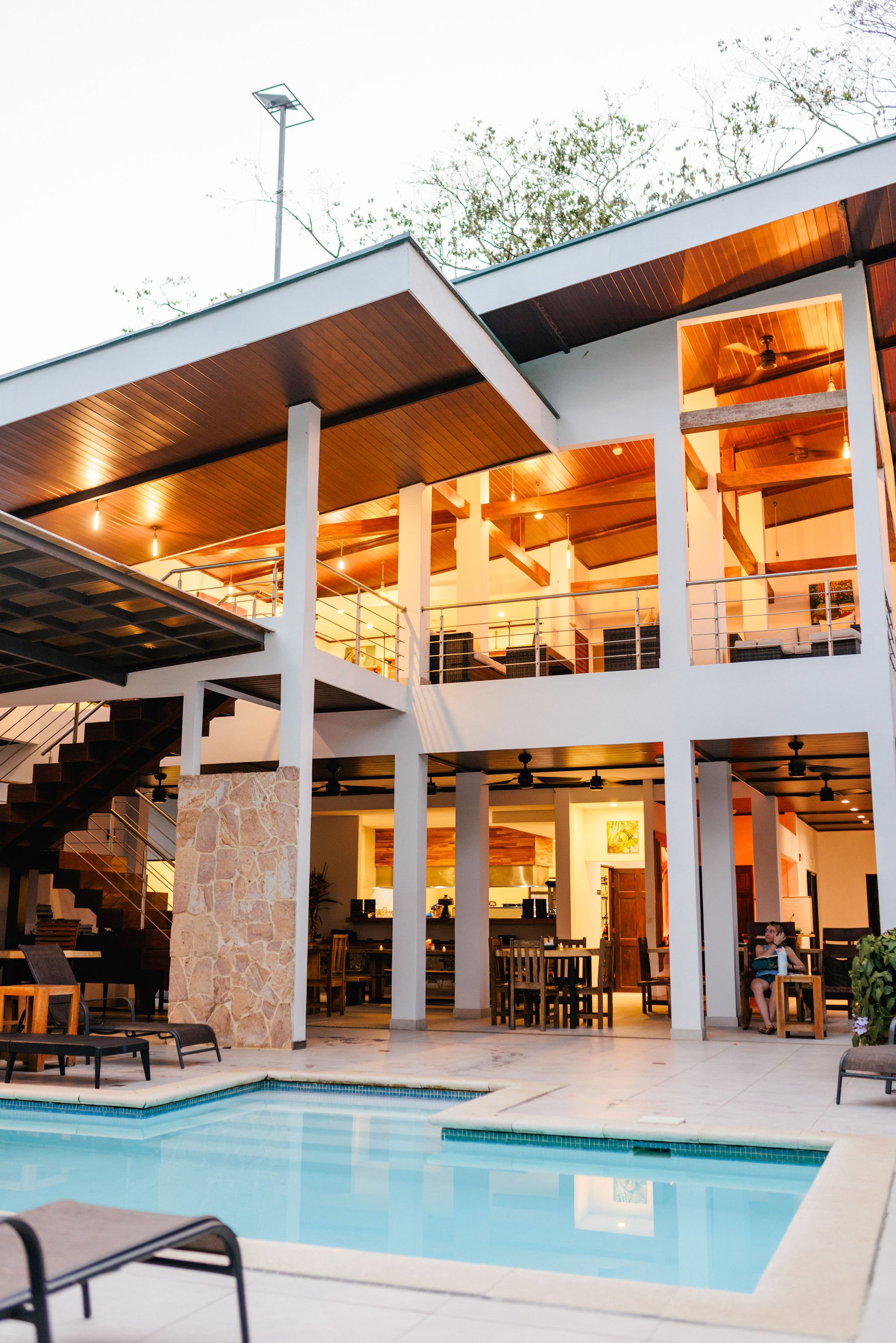 View of a modern, two-story house with a swimming pool in the backyard, illuminated softly inside during evening, with a woman sitting on a chair near the pool.