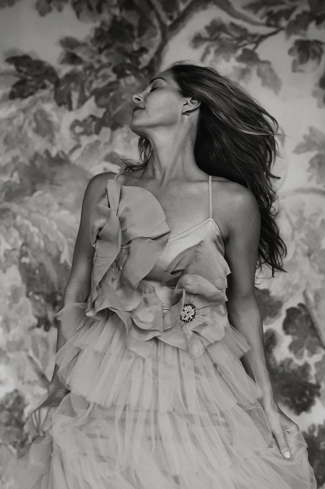 A woman with long flowing hair, wearing a strap dress with a ruffled skirt and large floral embellishment, posed with her head tilted back and eyes closed, in front of a floral background.
