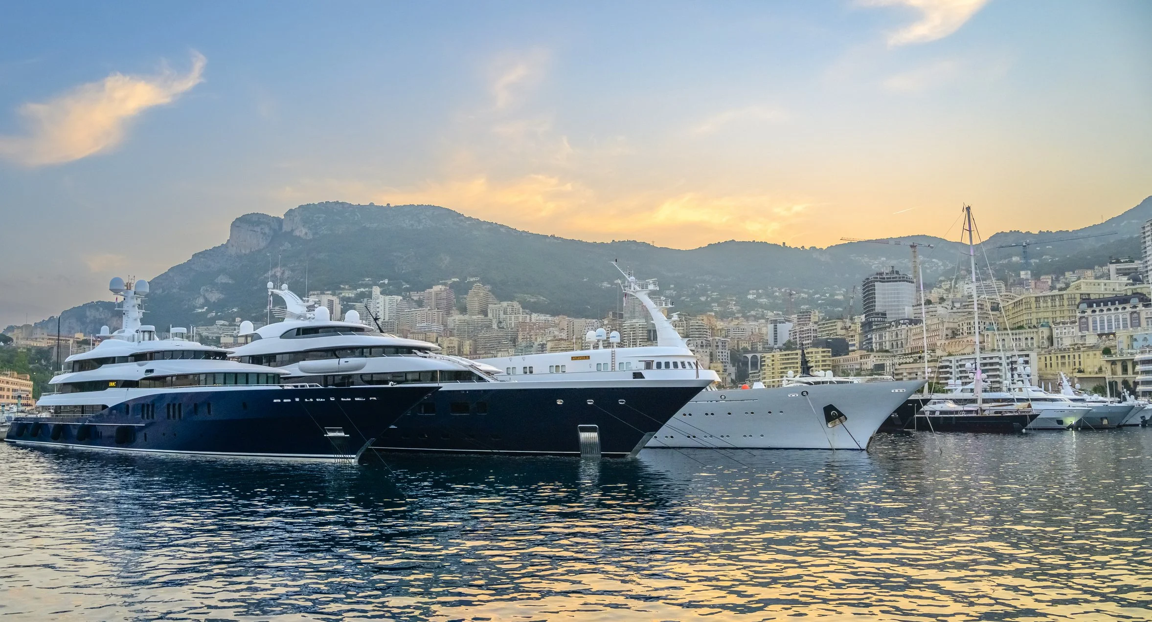 Luxury yachts docked in a marina with a cityscape, showcasing Over Oceans Yacht Interiors Projects & Management.