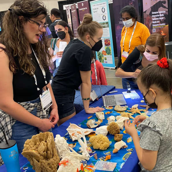 A group of people, including children and adults, wearing face masks, gathered around a table displaying various coral and marine life specimens, at an indoor science or education event.