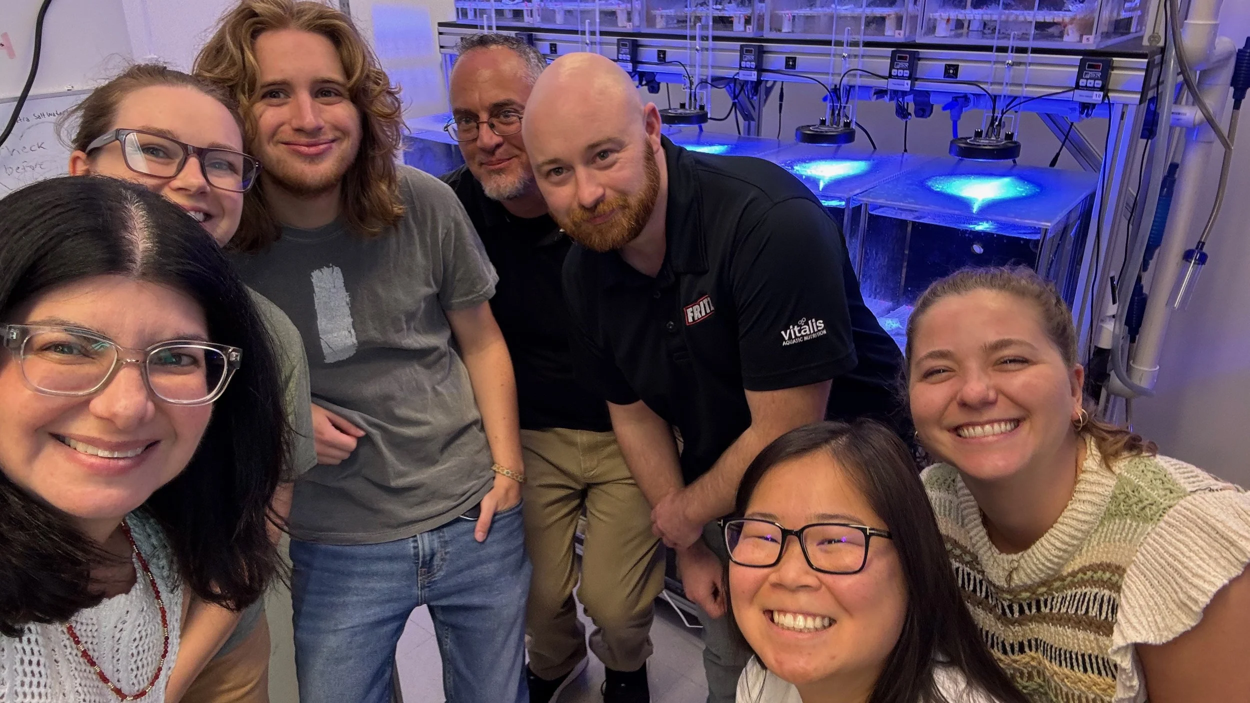 Group of diverse smiling people taking a selfie in a laboratory with scientific equipment and blue lighting in the background.
