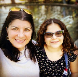 Two women outdoors in front of a fountain, smiling at the camera. One woman has dark hair and is wearing a white top, while the other has curly hair and sunglasses, wearing a black floral top.