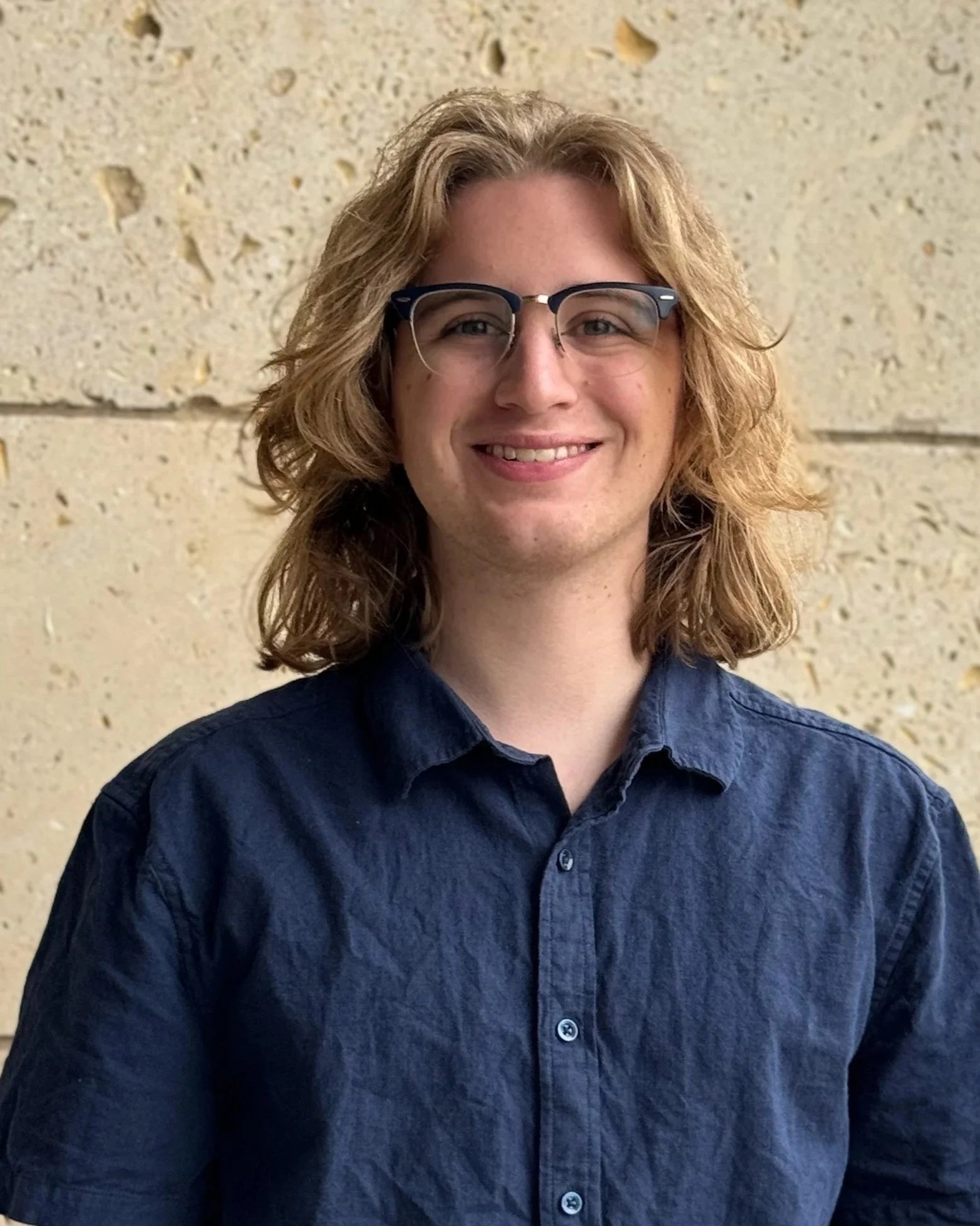 A person with curly long red hair and glasses, smiling, wearing a dark blue shirt, standing in front of a beige, textured wall.