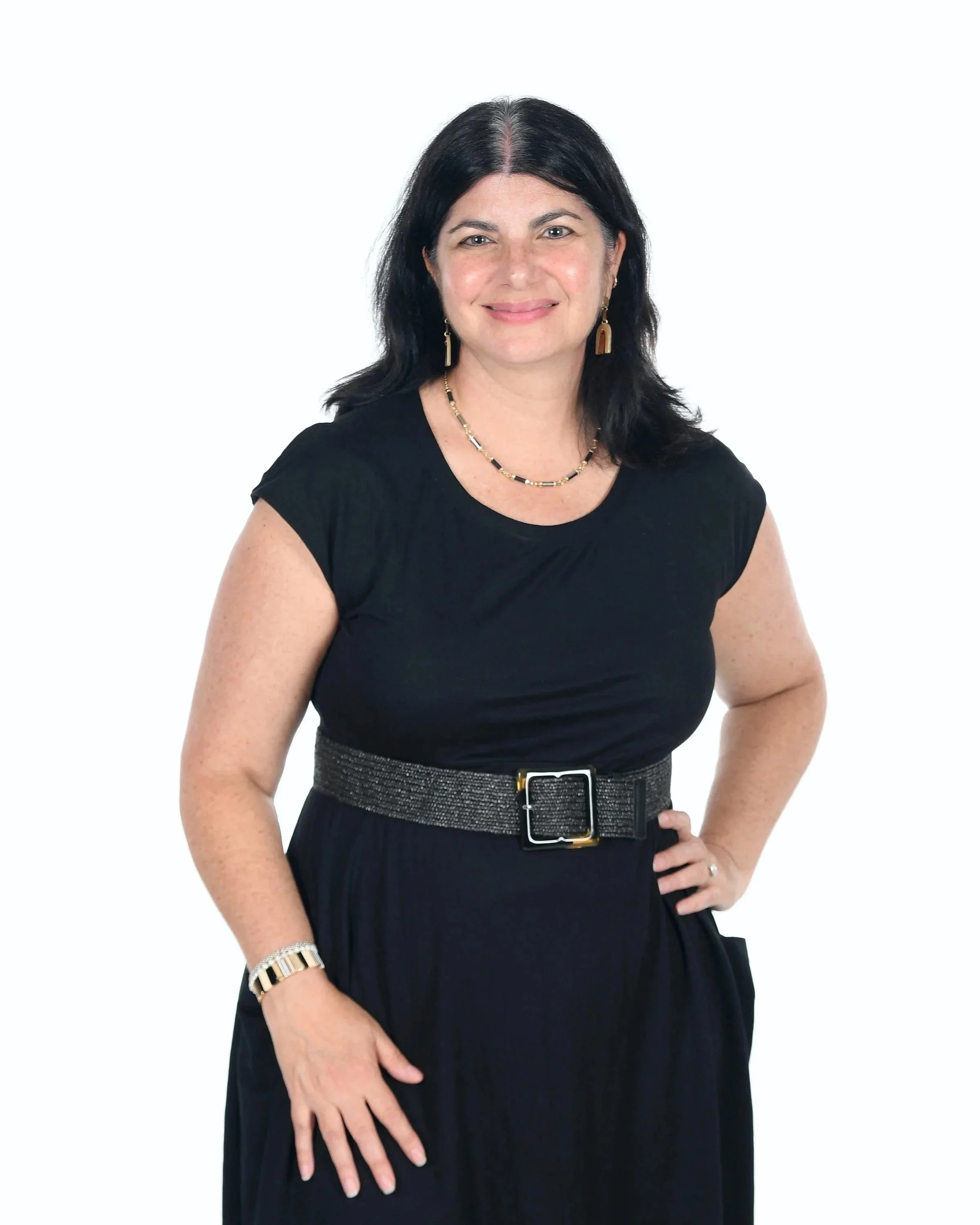 A woman with dark hair wearing a black top, gold jewelry, and a black belt, standing against a plain white background.
