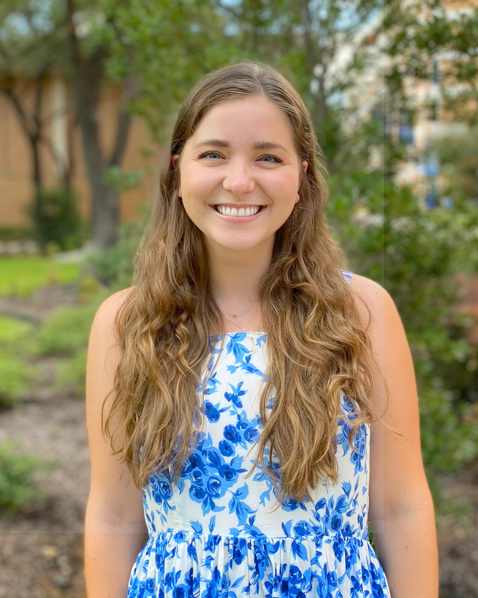 A young woman with curly long hair and blue eyes smiling outdoors, wearing a white dress with blue floral patterns, in a park with trees and buildings in the background.