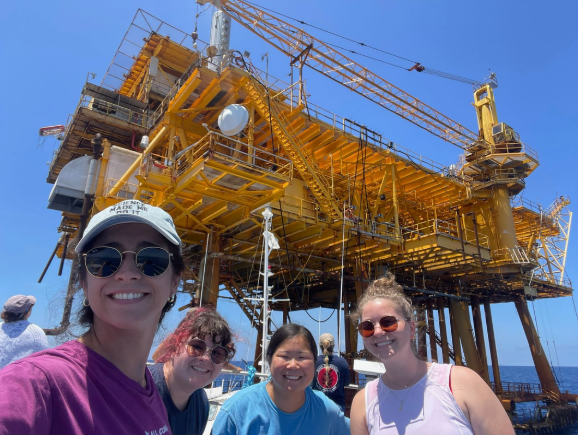 Four women taking a selfie on a boat with an offshore oil rig in the background.