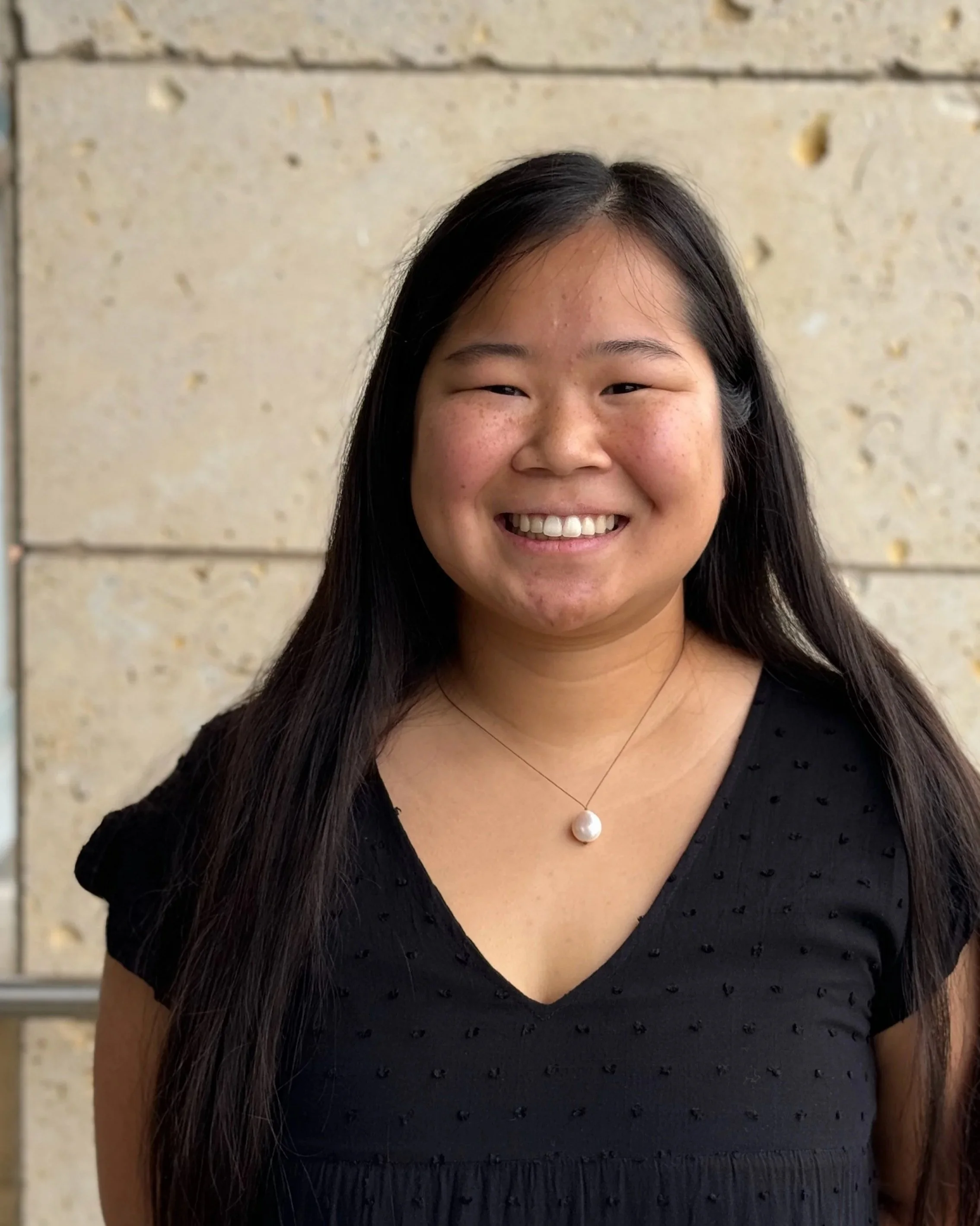 A woman with long dark hair smiling in front of a beige stone wall. She is wearing a black dress with small textured details, a necklace with a large pearl pendant, and has makeup with blush and lipstick.