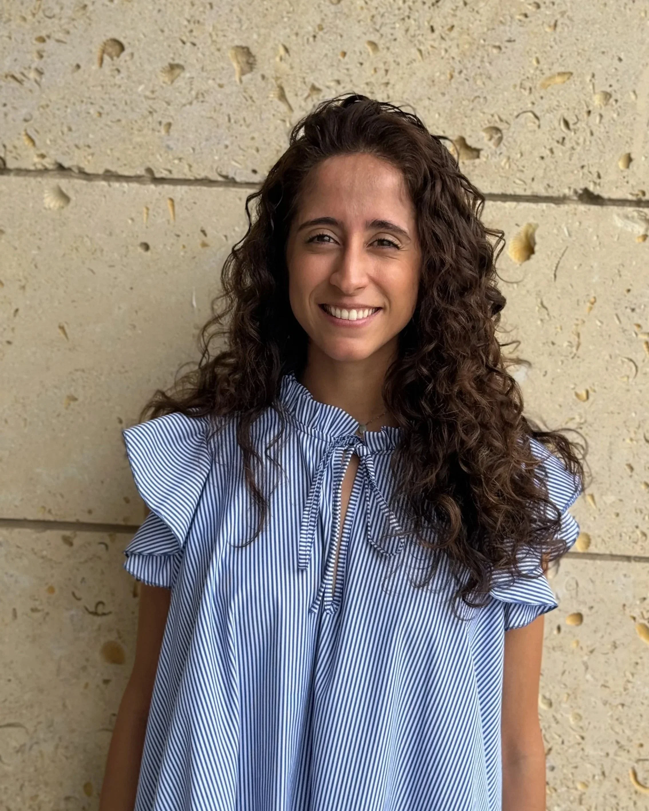 A woman with curly hair, wearing a blue and white striped blouse, smiling, standing against a beige, textured wall.