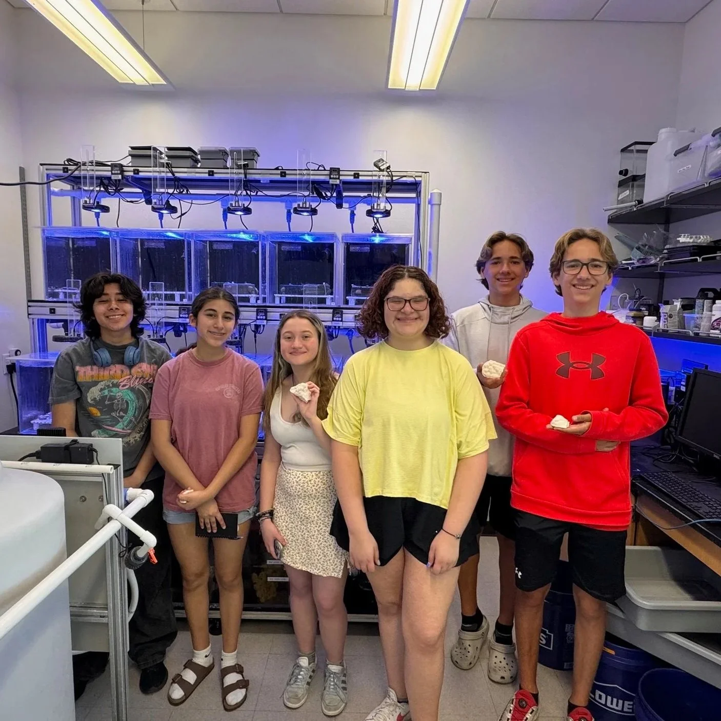 Group of six children standing in a laboratory with scientific equipment and tanks in the background, holding small objects.
