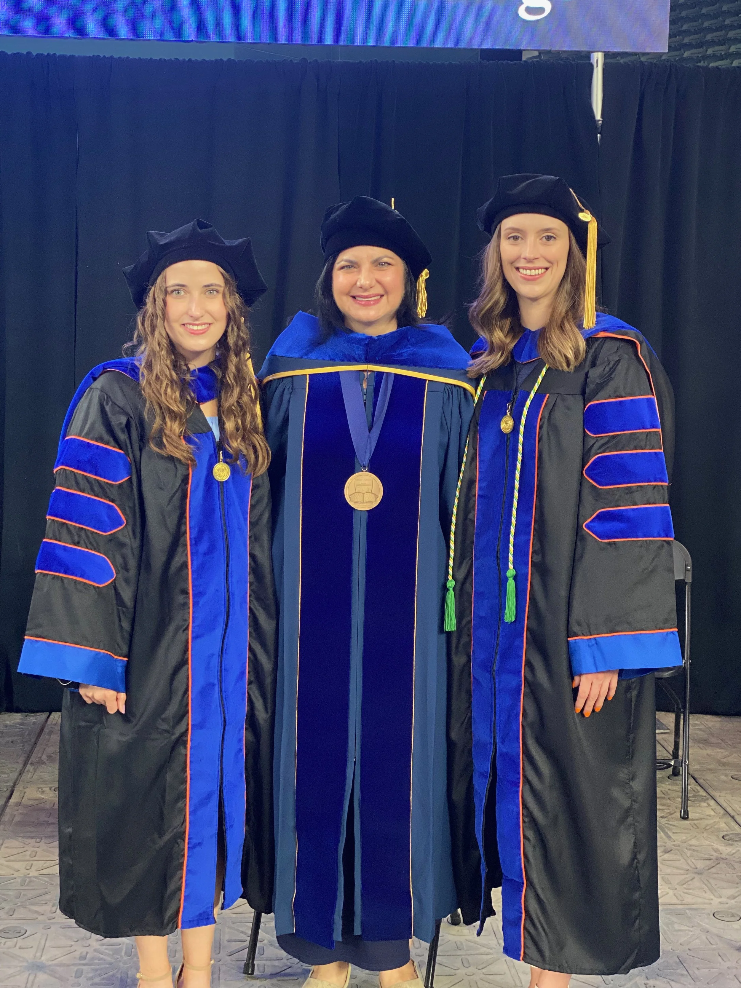 Three women dressed in academic regalia, including gowns and caps, standing together at a graduation ceremony. The woman in the middle is wearing a black gown with blue accents and has a gold medallion around her neck, indicating she is likely a faculty member or speaker.