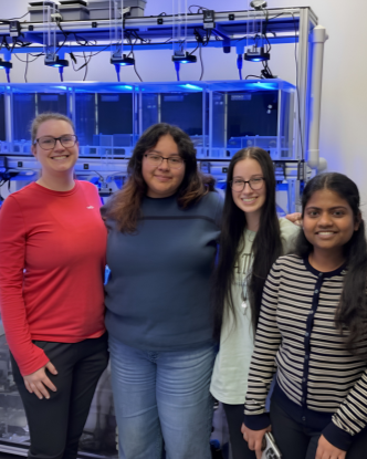 Four women standing in front of scientific equipment with illuminated containers and lab apparatus in a laboratory setting.