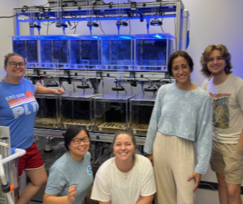 Five women in a laboratory with fish tanks and equipment, smiling and posing for the photo.