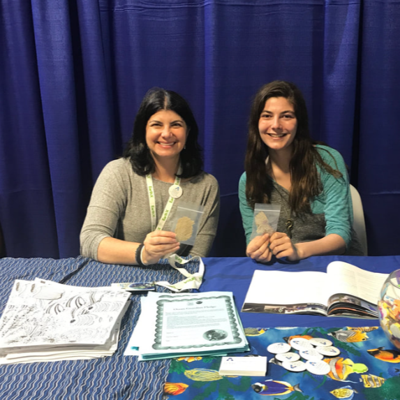 Two women smiling and sitting at a table with books, papers, and lanyards, in front of a blue curtain.