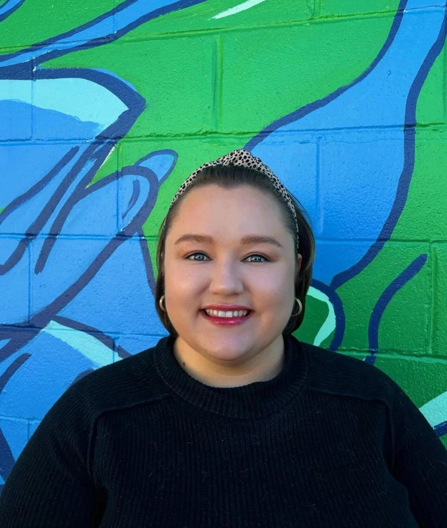 Close-up of a smiling woman with short dark hair, wearing a black sweater and a polka dot headband, standing in front of a colorful graffiti wall with blue, green, and white abstract designs.