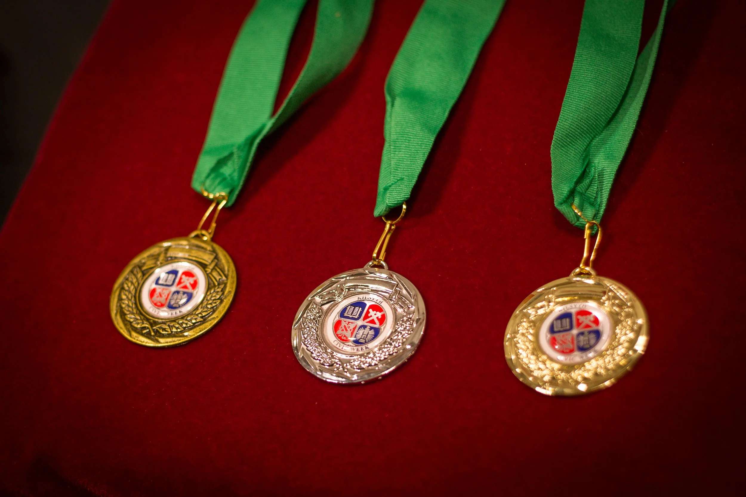 Three medals with green ribbons arranged on a red surface, each medal featuring a circular emblem with a shield divided into four sections with books and a torch.