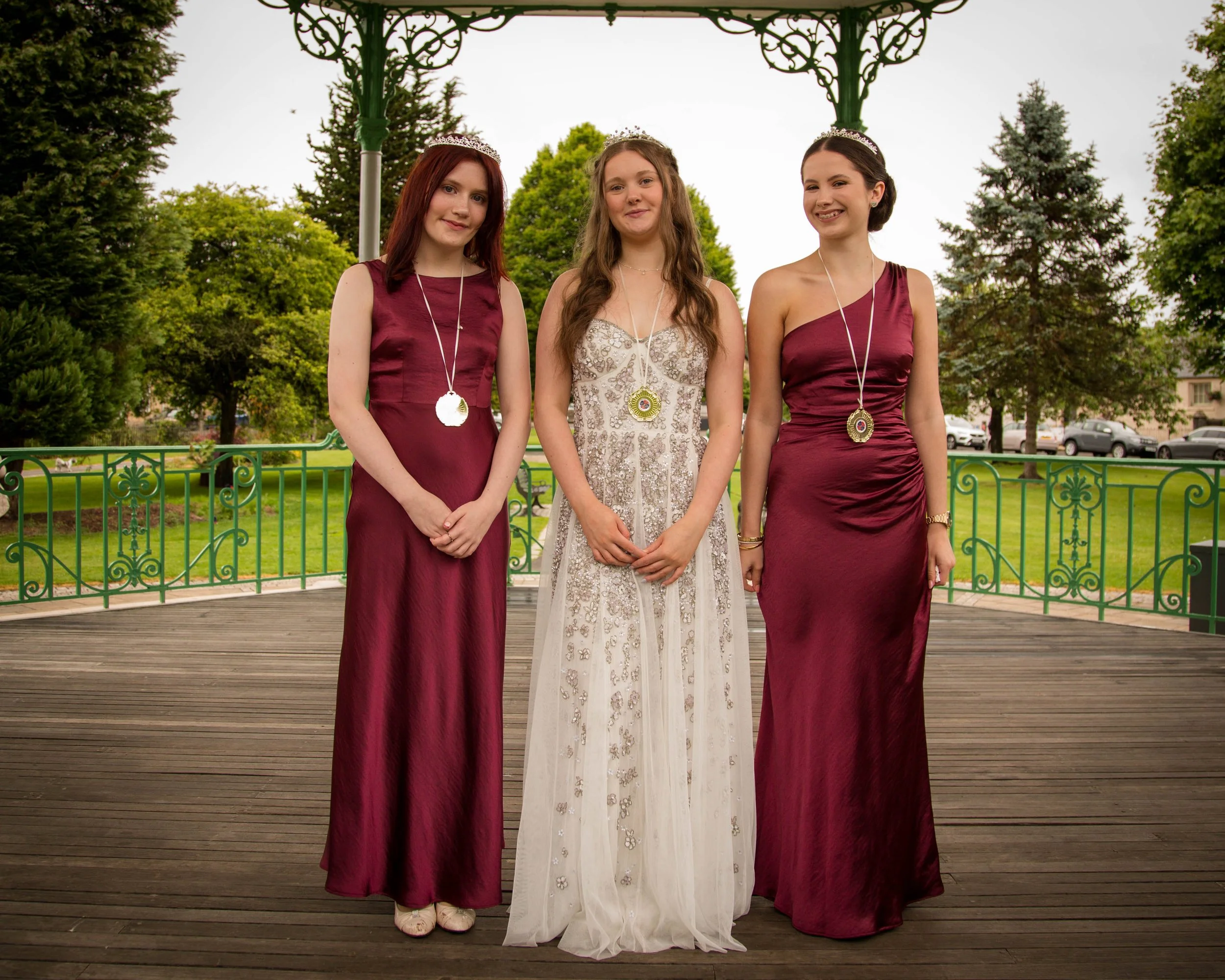 Three young women dressed in formal gowns, standing on a wooden platform outdoors, wearing tiaras and medals around their necks, with trees and parked cars in the background.