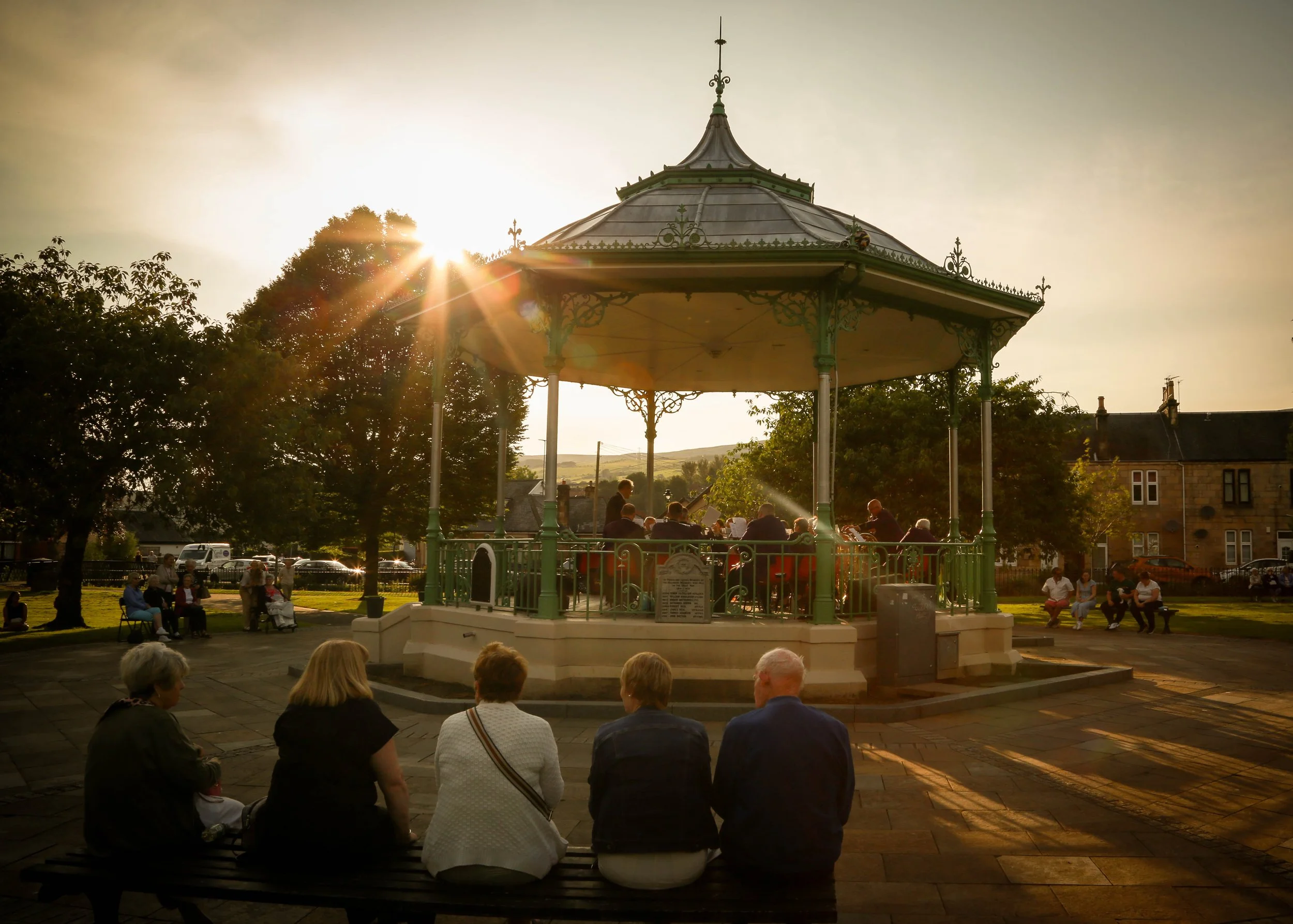 A band playing in a gazebo in a park at sunset, with people sitting on benches and standing around watching.