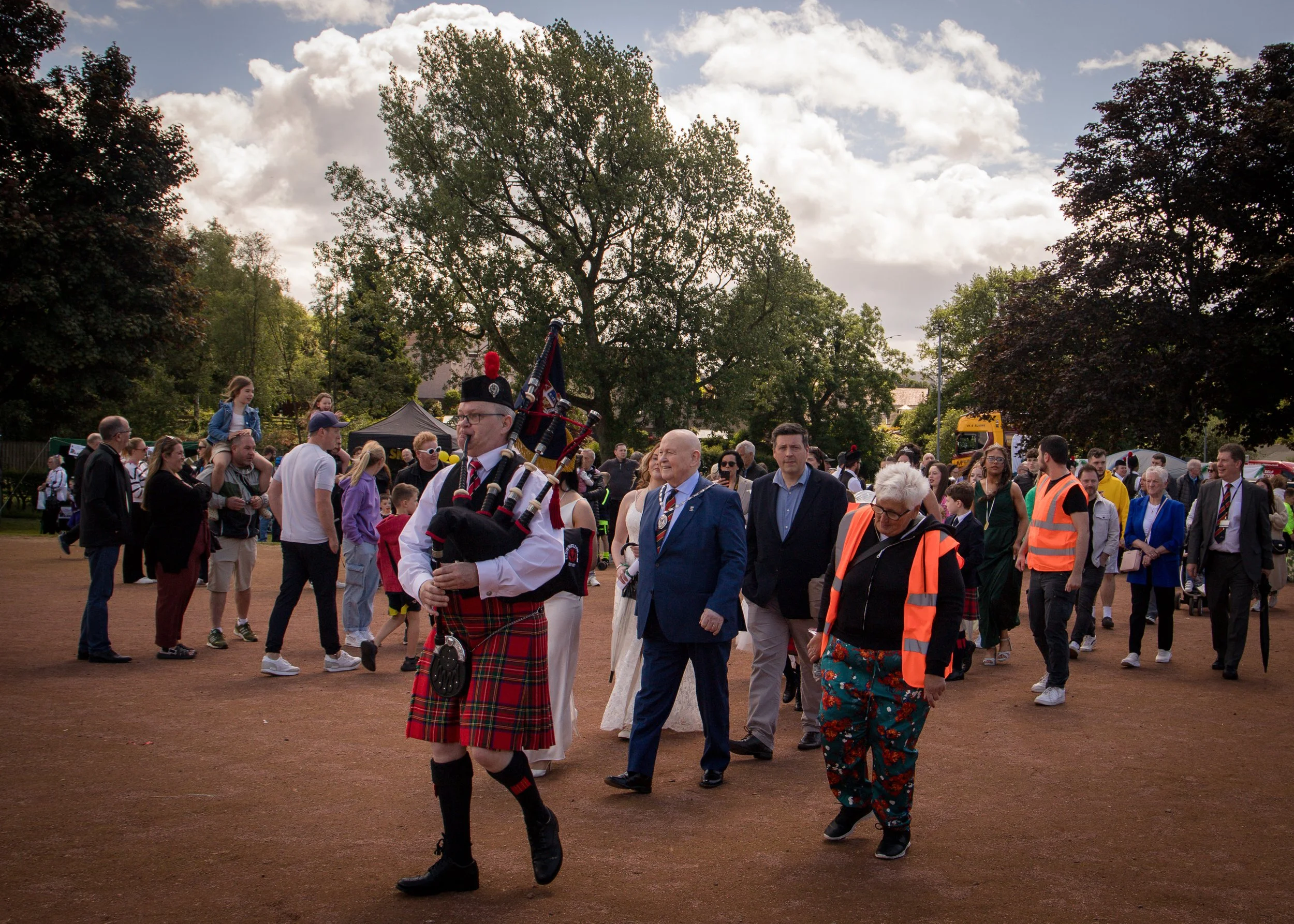 A group of people walking outdoors on a dirt path during the daytime, with one man in a traditional Scottish kilt playing bagpipes at the front. The crowd includes individuals of various ages, some in formal attire and some in casual clothing, with a background of trees and a partly cloudy sky.