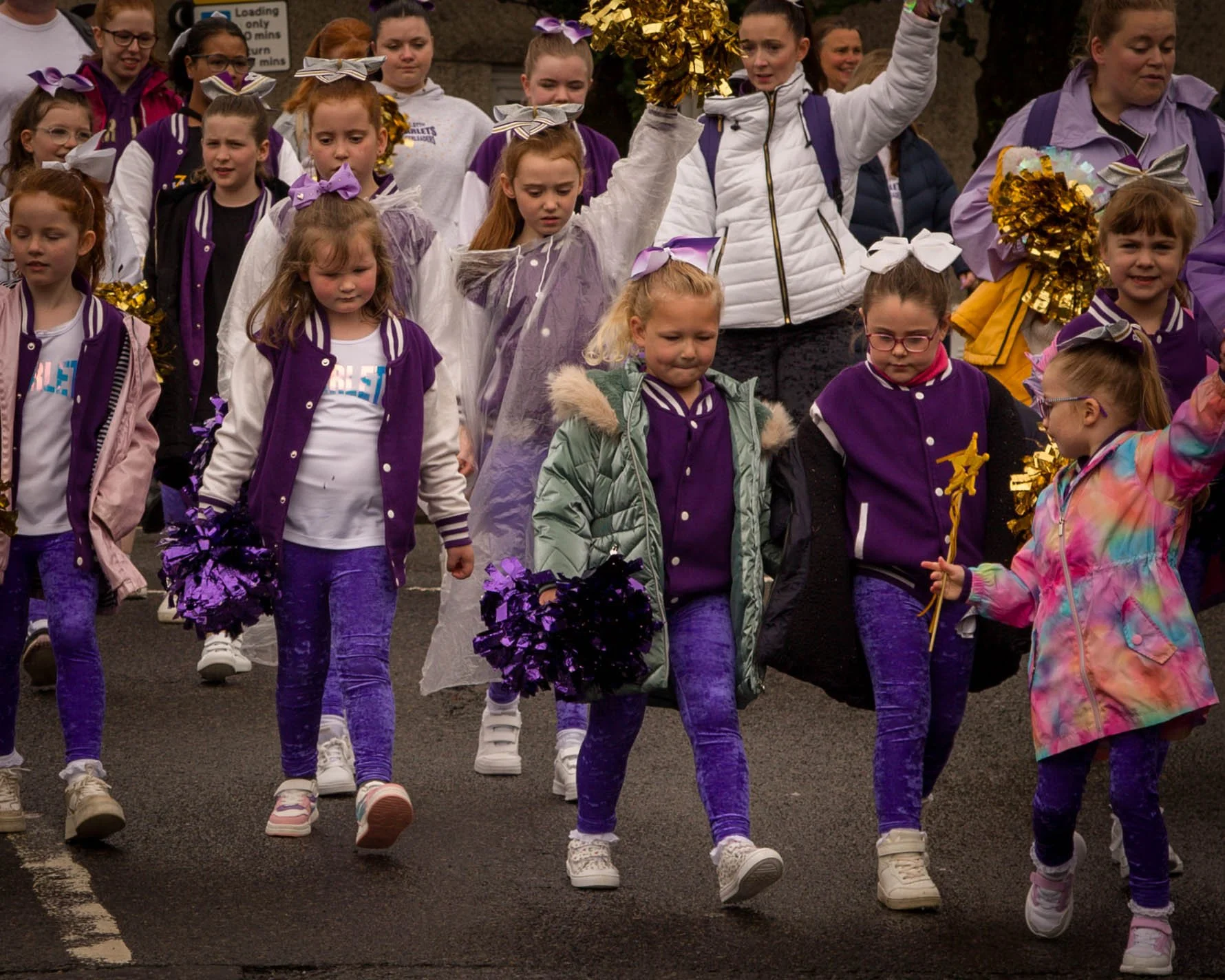 A group of young girls participating in a parade, dressed in purple and white cheer leading uniforms, carrying purple and gold pom-poms, walking on a paved road.