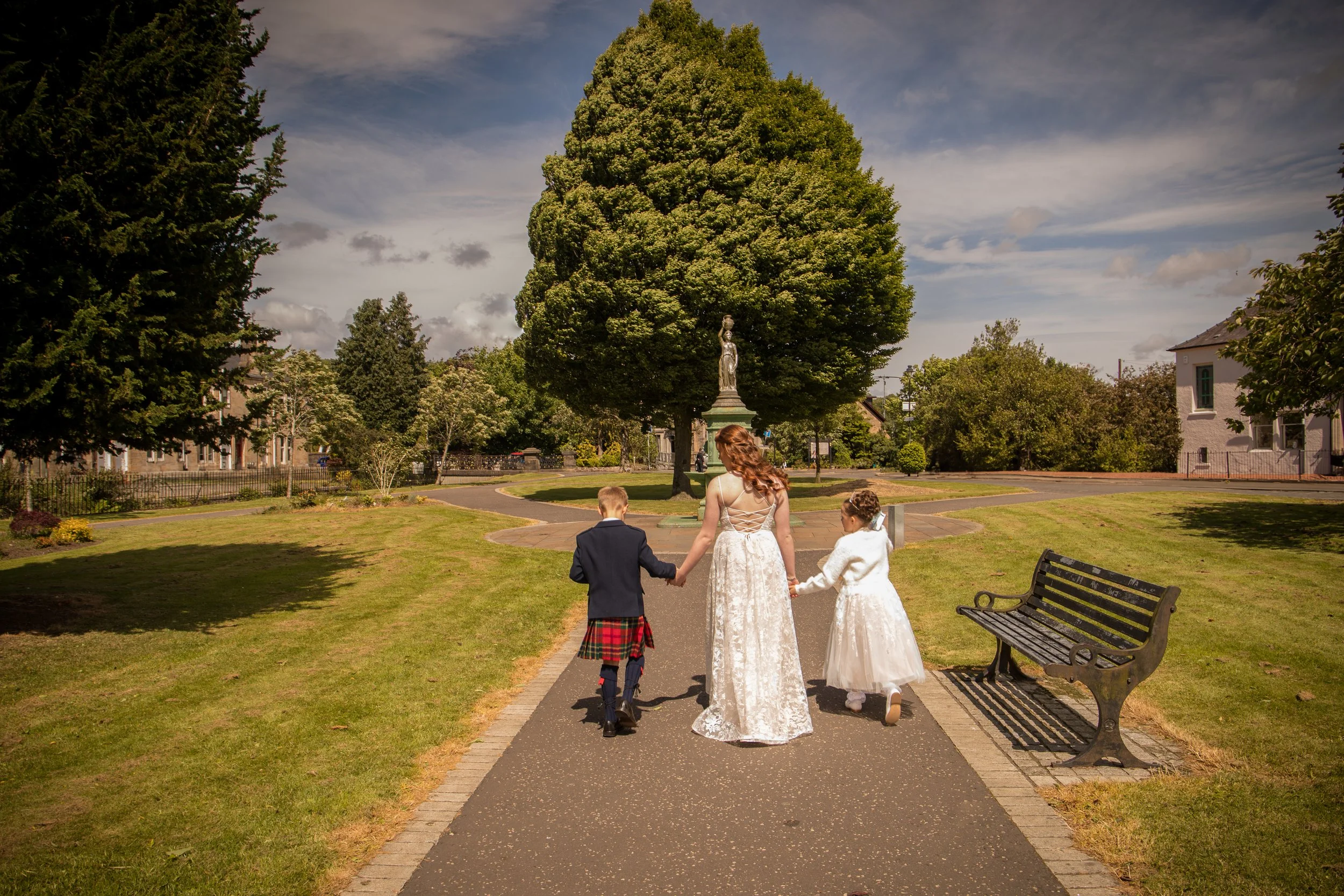 A woman in a white lace dress walking hand-in-hand with two children, a boy in a kilt and a girl in a white dress, through a park with green grass and trees, on a sunny day.