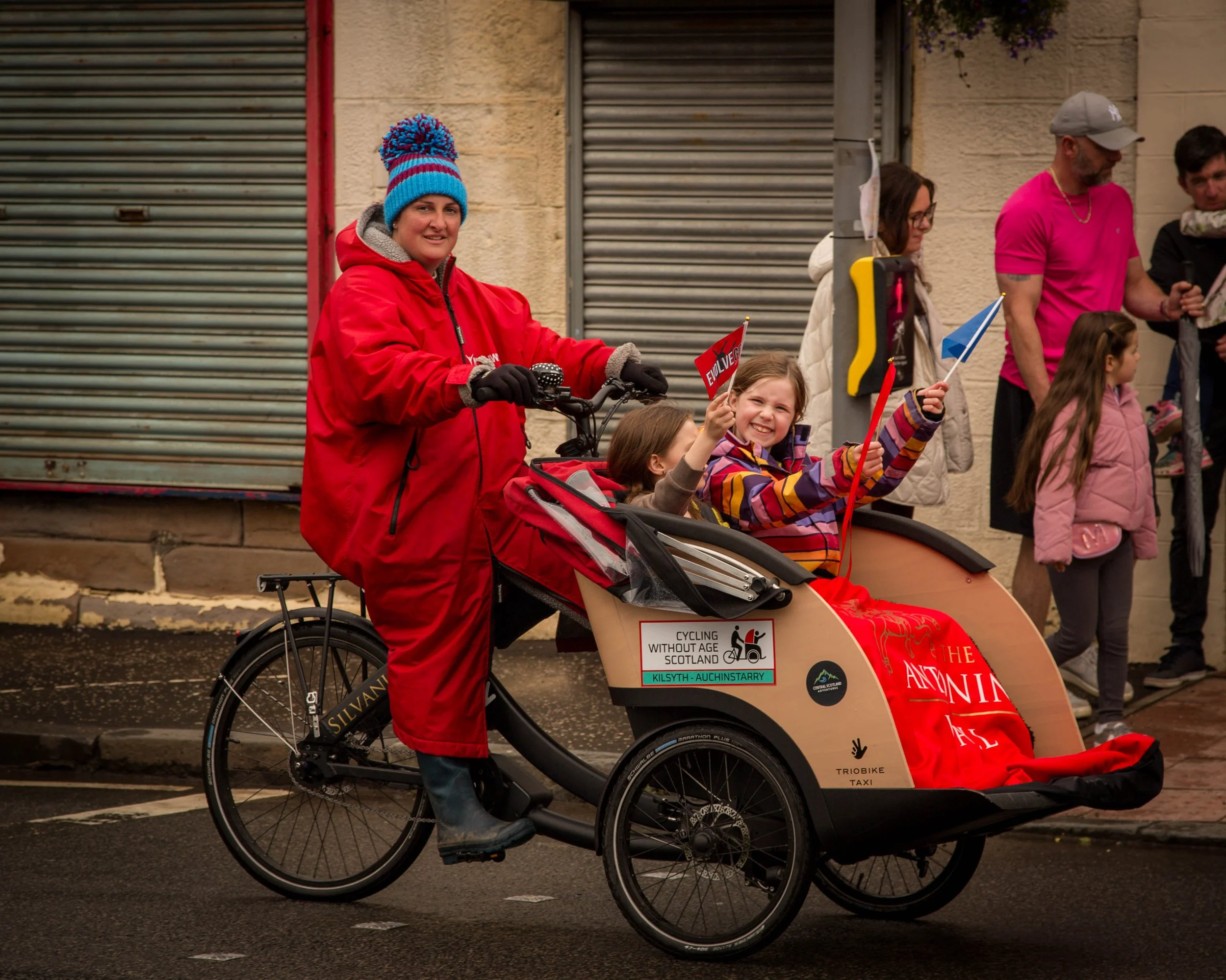 A woman in a red jacket and blue knit hat rides a tricycle with two young girls seated inside, holding small flags, during a parade.