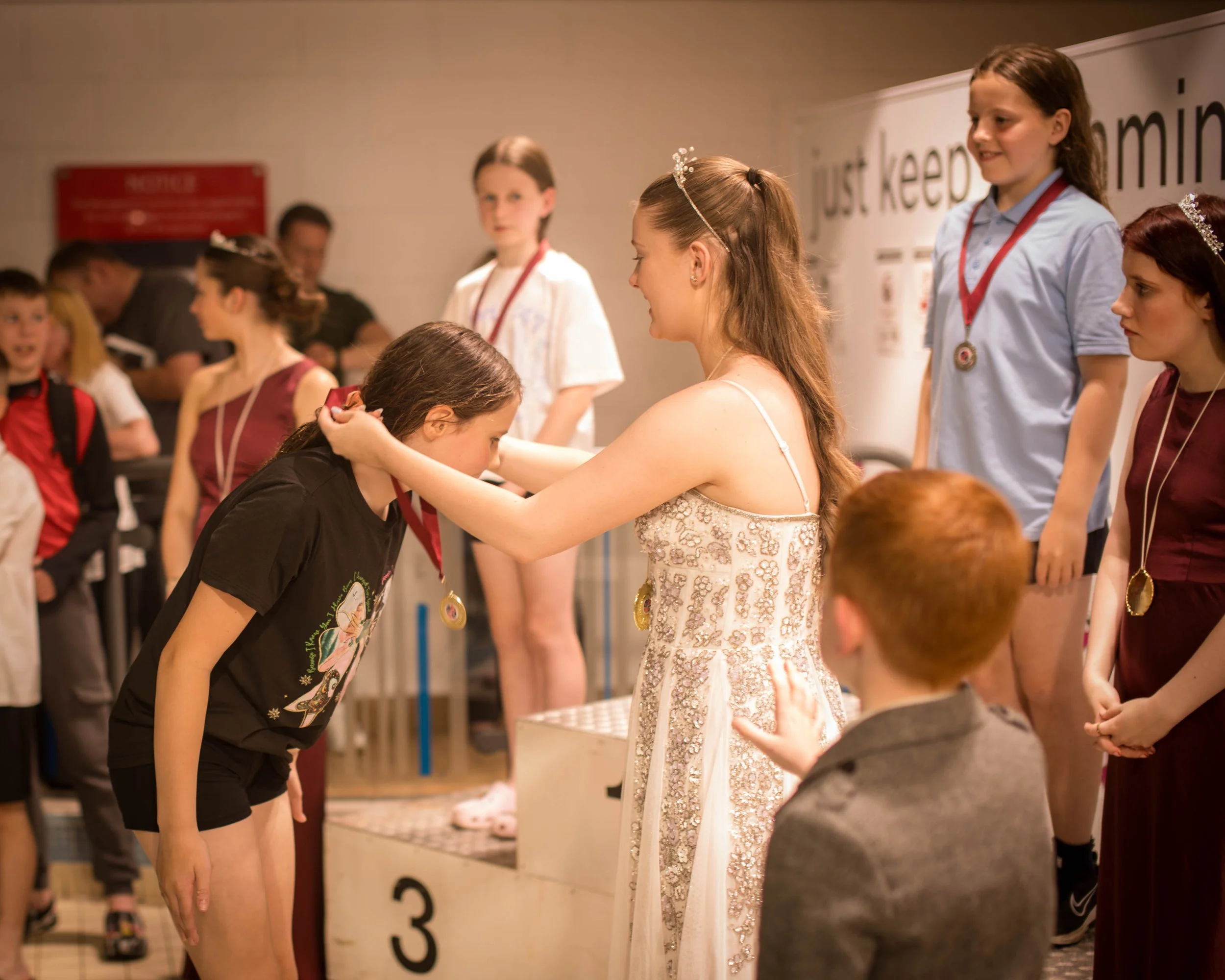 A girl receiving a medal at an award ceremony as others wait their turn, with children wearing medals on a podium and a background sign that reads "just keep dreaming."