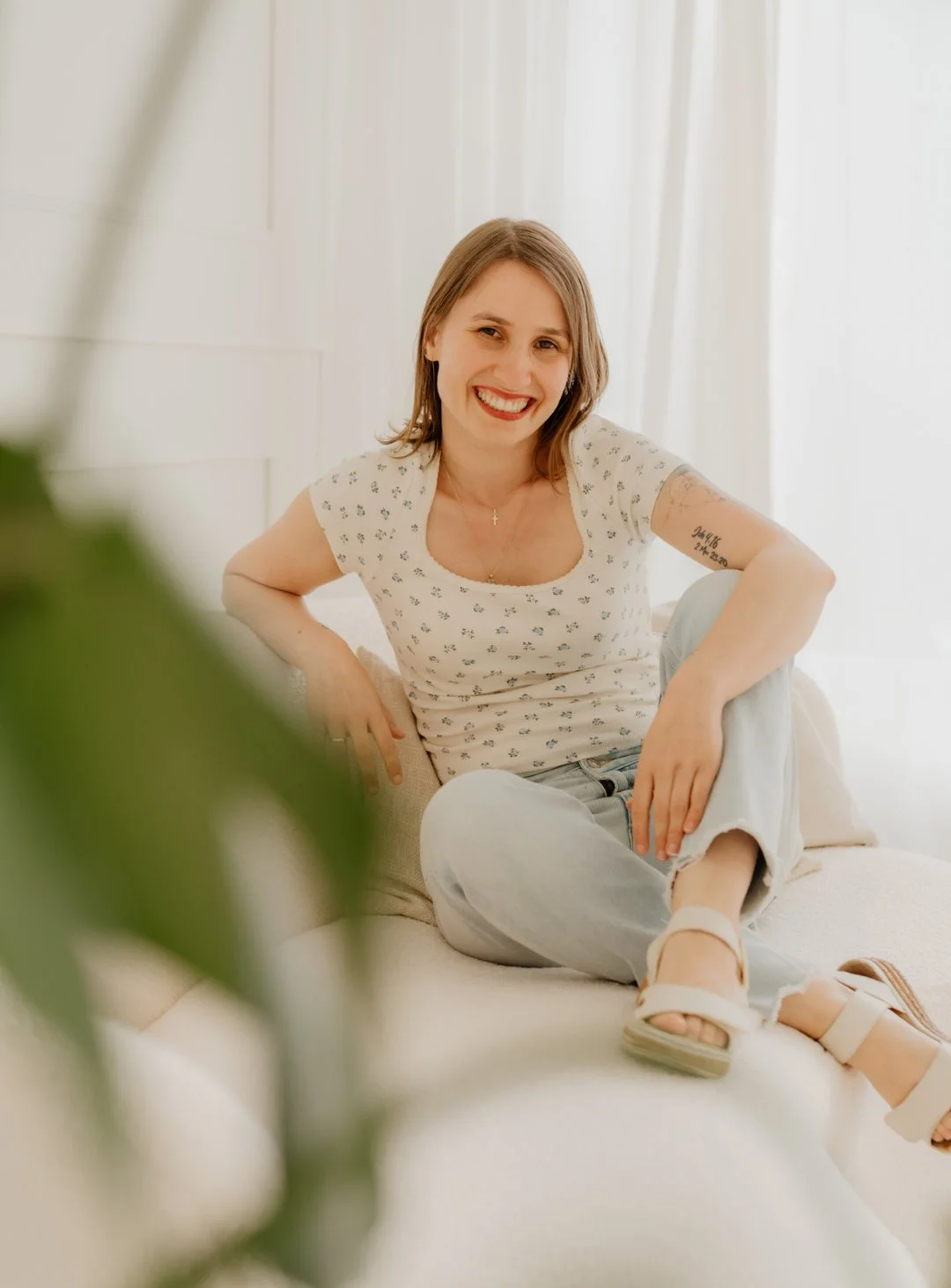 Die Künstlerin sitzt auf einem Sofa und lächelt in die Kamera, der Blickwinkel erfolgt durch Blätter einer Pflanze