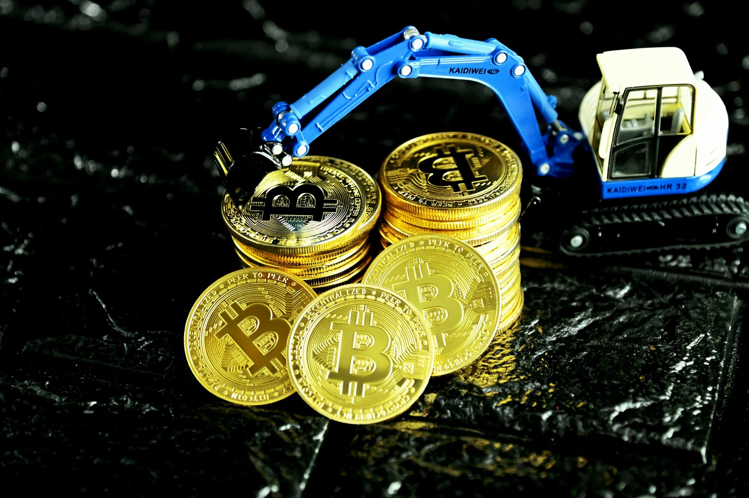 Stack of gold Bitcoin coins with miniature construction excavator toy placed on top, on a dark textured surface.