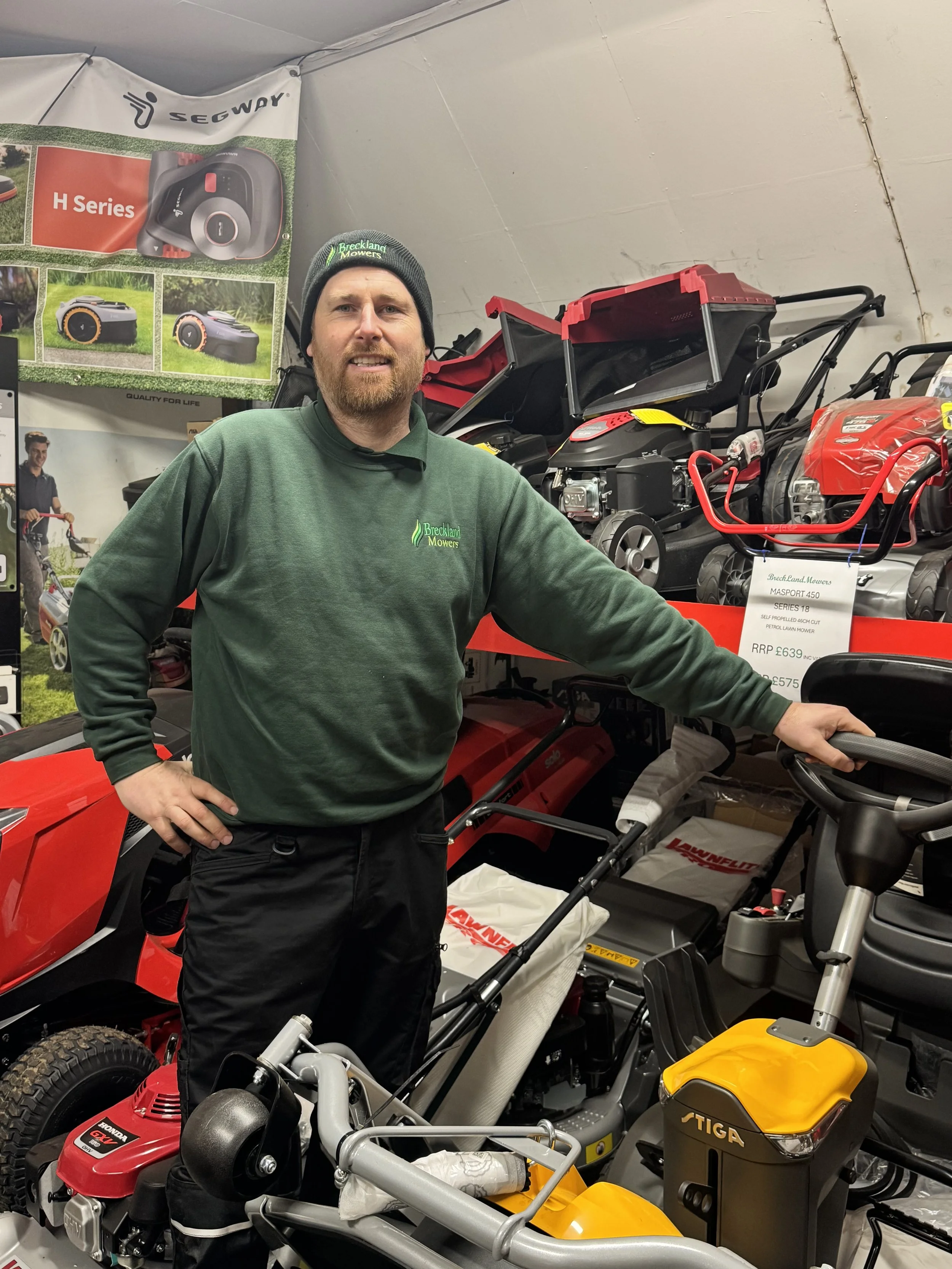 Man posing indoors at a display of outdoor power equipment and vehicles, including lawnmowers, and multiple power machines, with promotional posters in the background.