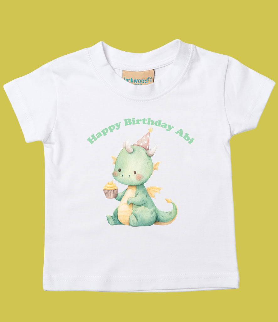 White children's T-shirt with a cute illustration of a green baby dragon wearing a pink party hat, holding a cupcake, with the text "Happy Birthday Abi" in green above.