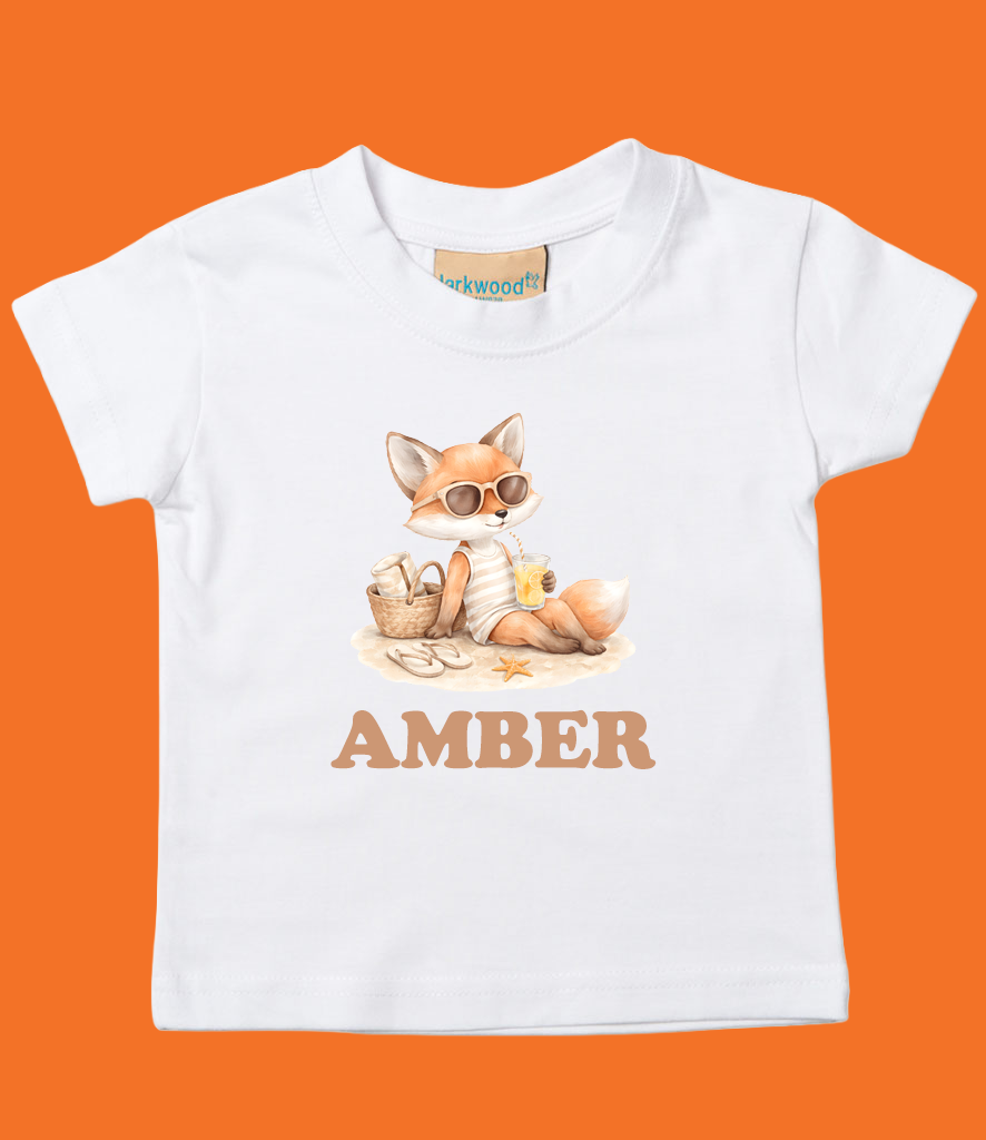 White toddler T-shirt with a cartoon fox wearing sunglasses, sitting on a beach, drinking a beverage with a straw, and surrounded by beach accessories. The name 'AMBER' is printed below the illustration.