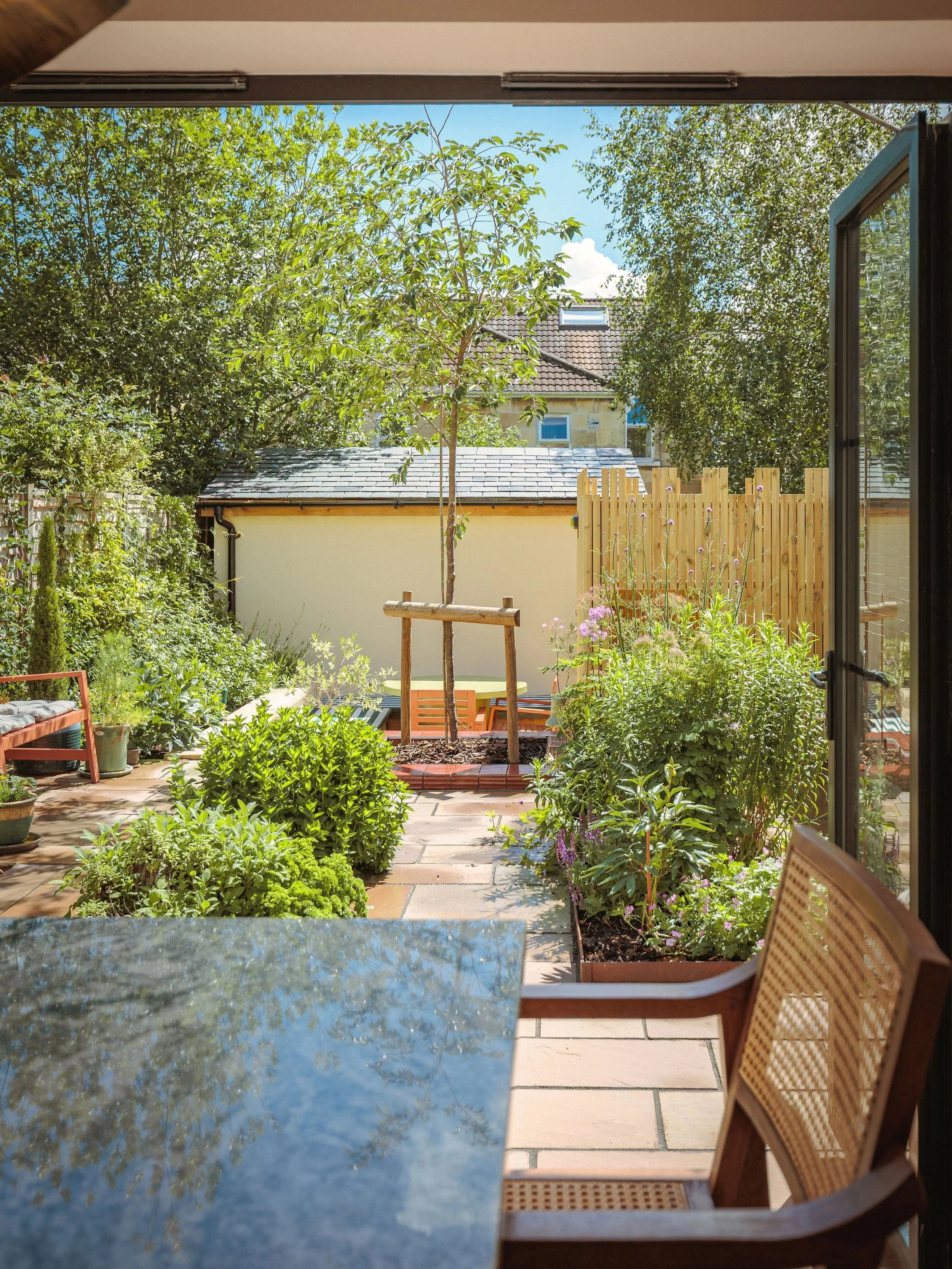 View from inside a house looking out onto a small city garden with various green plants, flowers, a young cherry tree, a wooden fence, a bespoke climbing frame and a house with roof skylights under a blue sky.