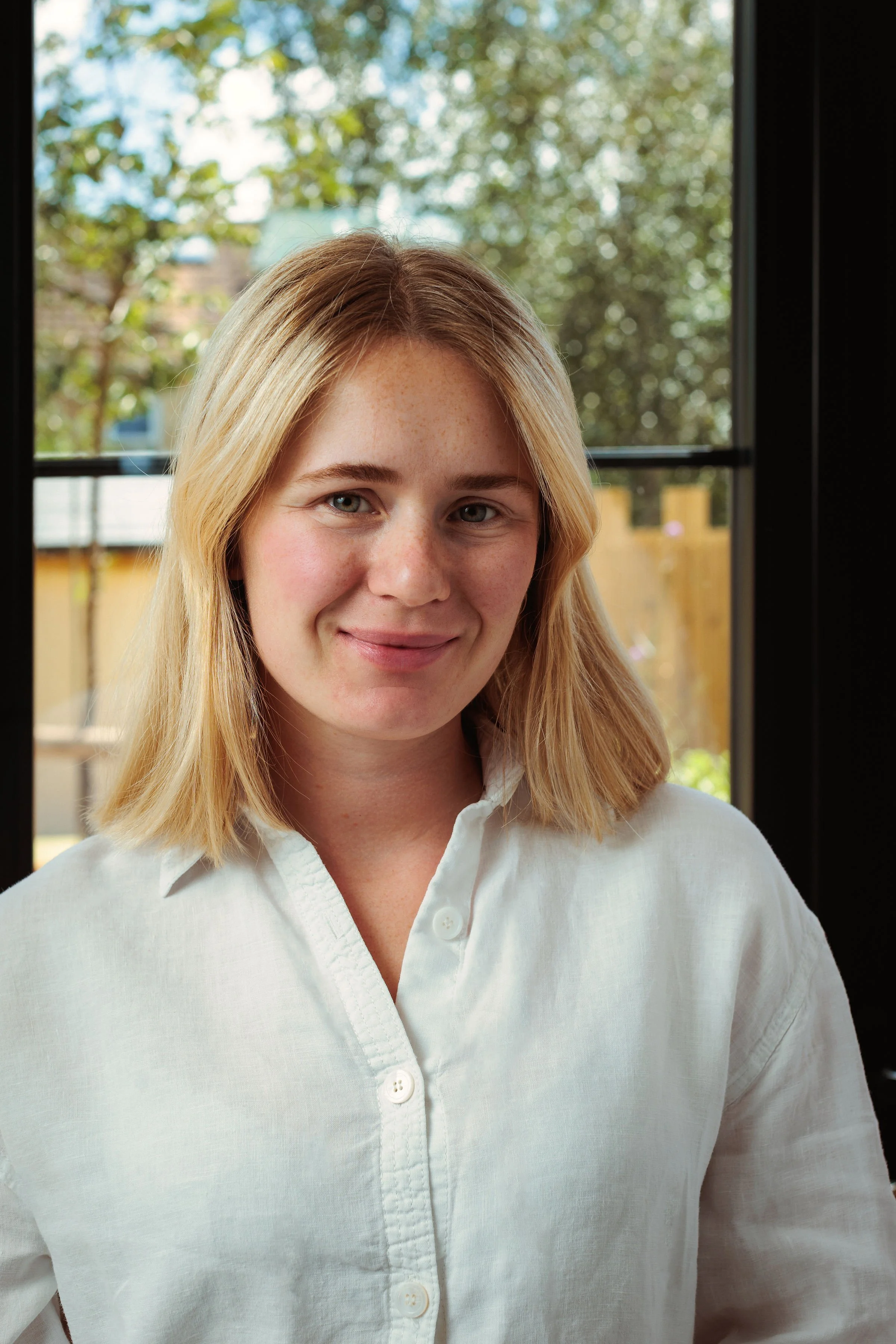 A young woman with blonde hair and green eyes, wearing a white button-up shirt, standing indoors near a window with greenery outside.