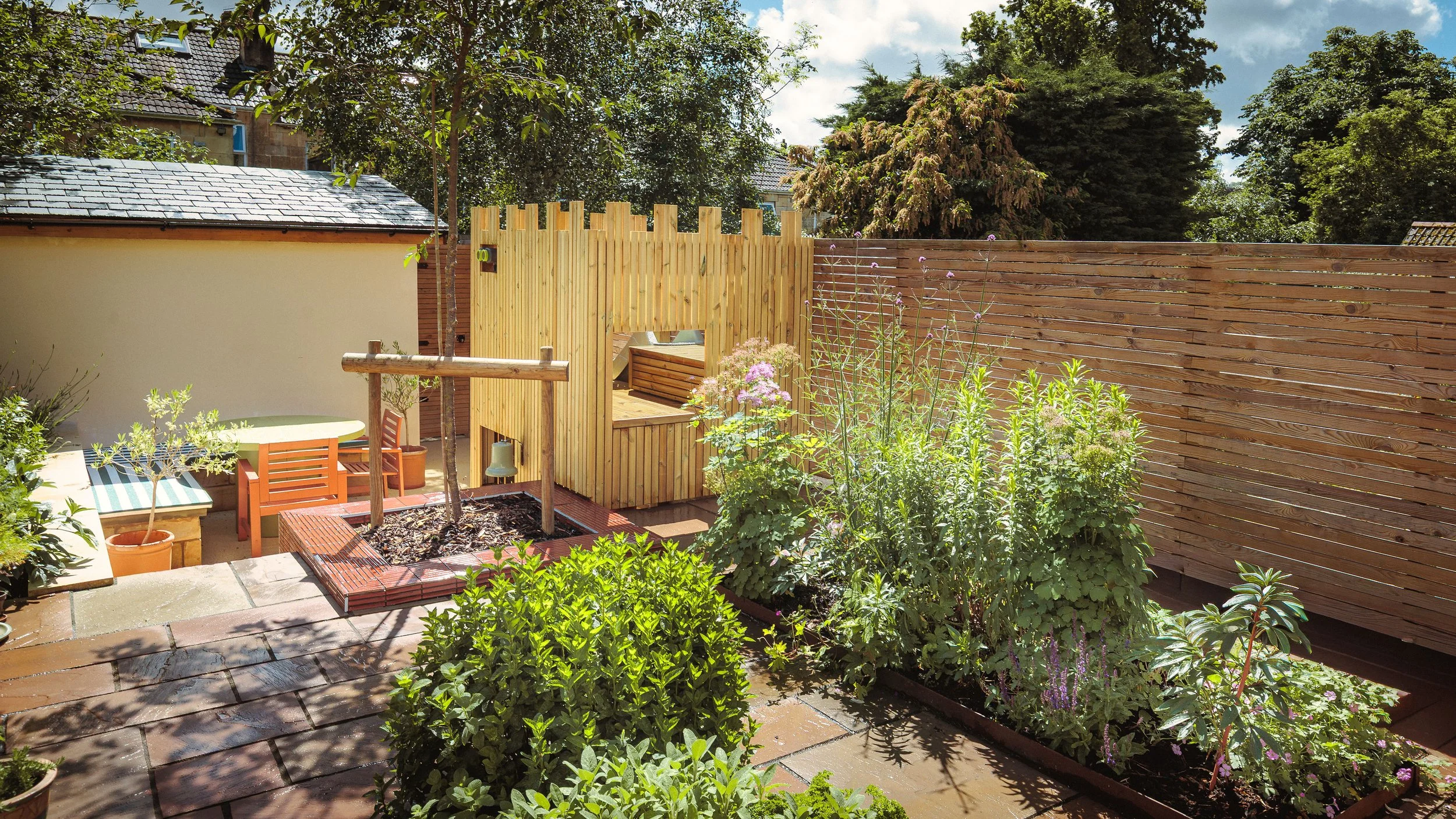 A backyard garden with a wooden fence, patio furniture, potted plants, and lush greenery under a sunny sky.