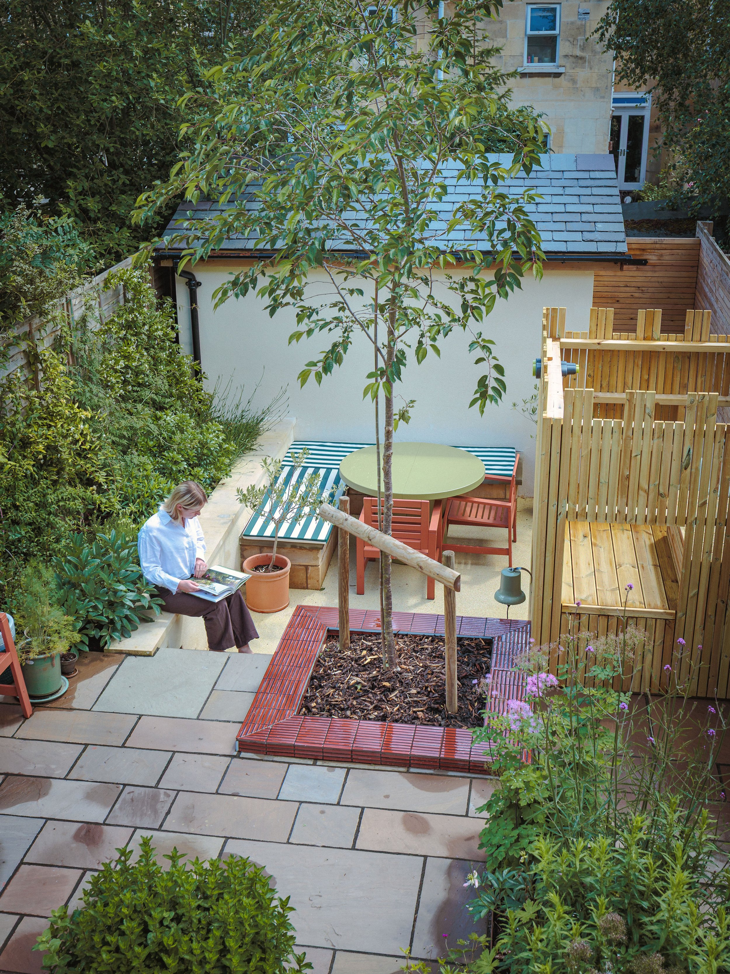 A woman sitting on a concrete bench in a garden, reading a magazine, surrounded by potted plants, trees, and a tiled patio. There is a small tree in the center enclosed by a brick border, and a wooden fence on the right. In the background, there is a