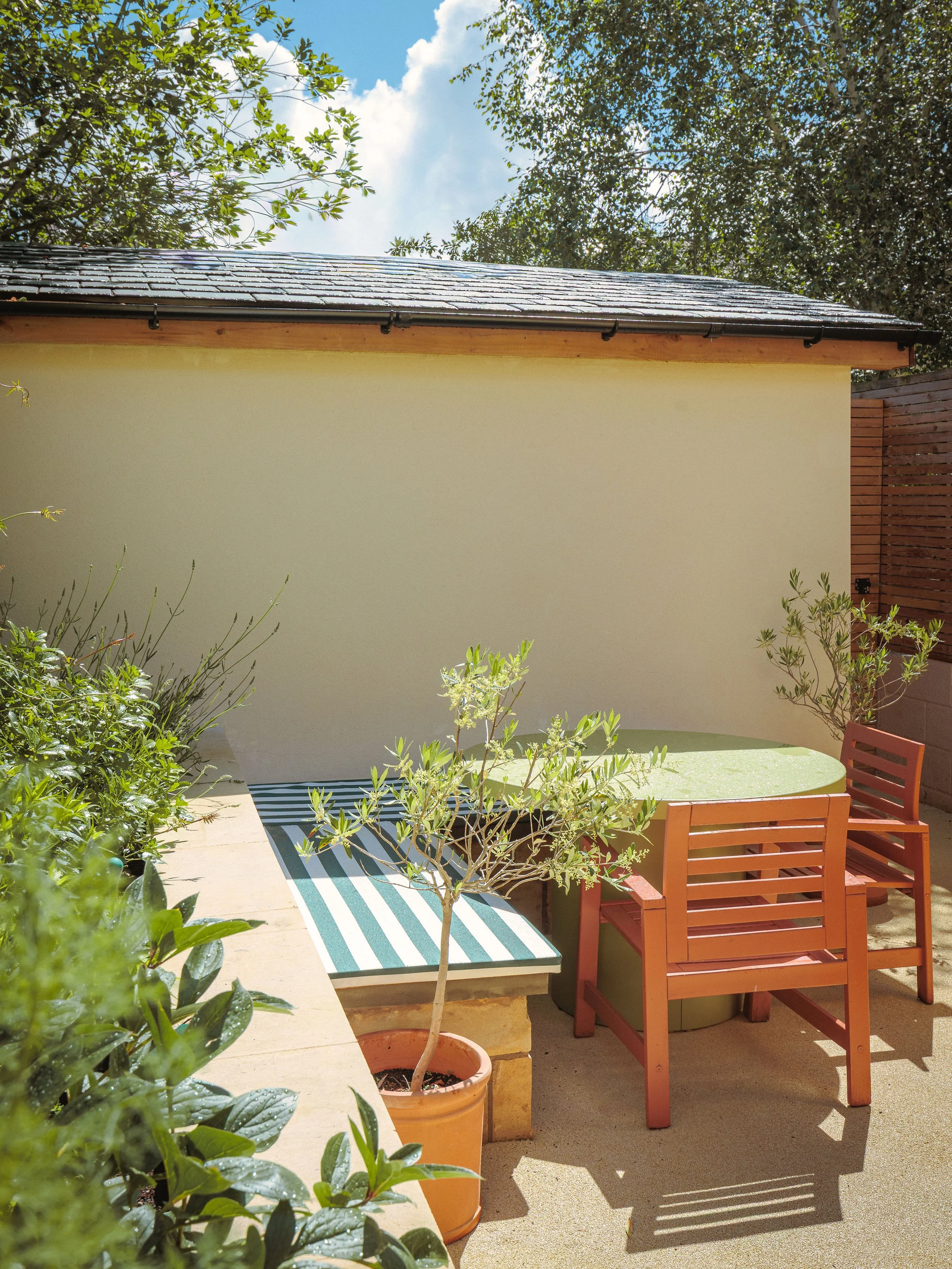 A small outdoor patio with a green round table and two wooden chairs, potted plants along the wall, a striped outdoor rug, and a fence with trees and a blue sky in the background.