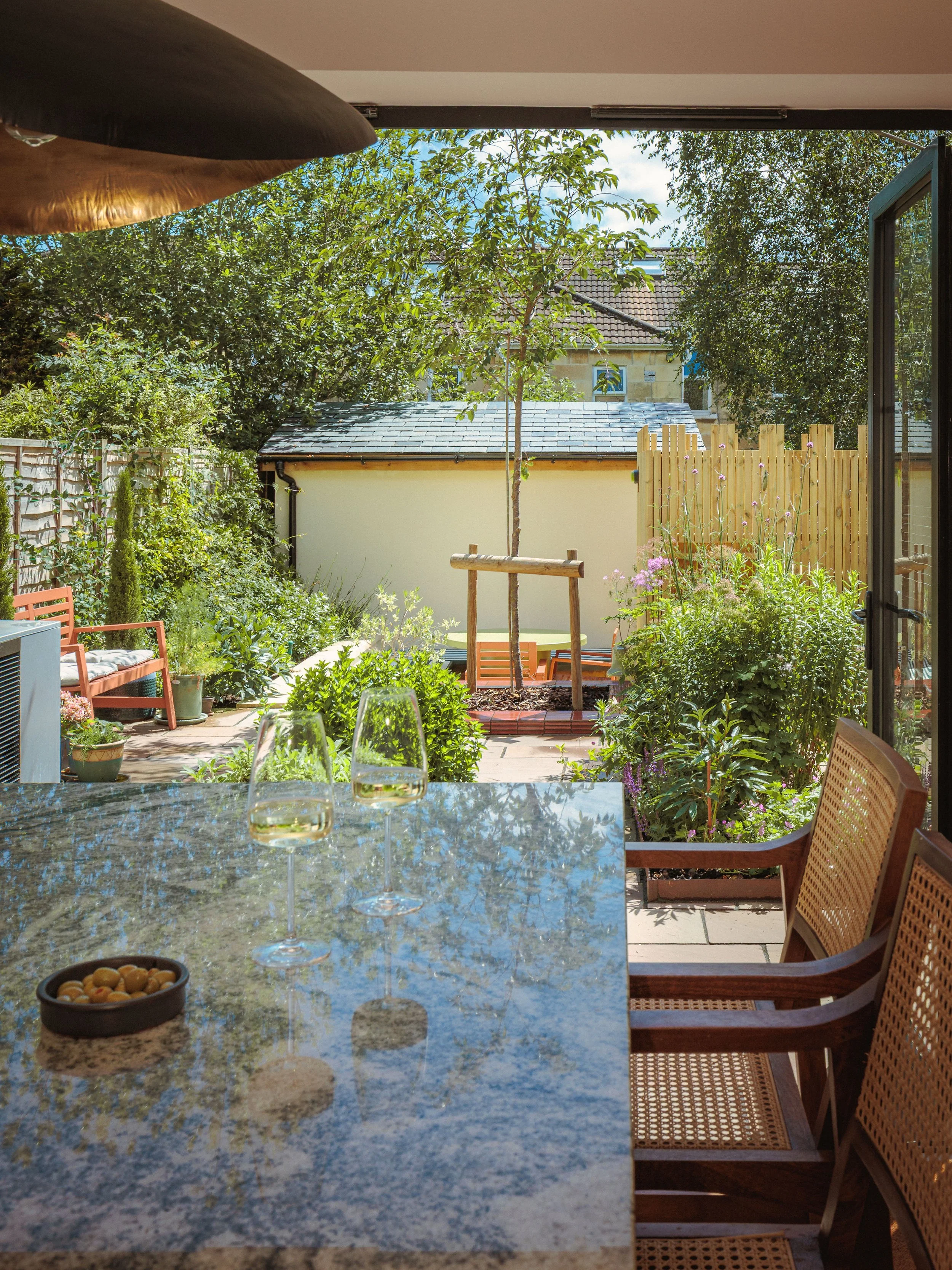 A view from inside a house looking out onto a patio with garden furniture, plants, and trees, and a small building with a tiled roof and a wooden fence in the background.